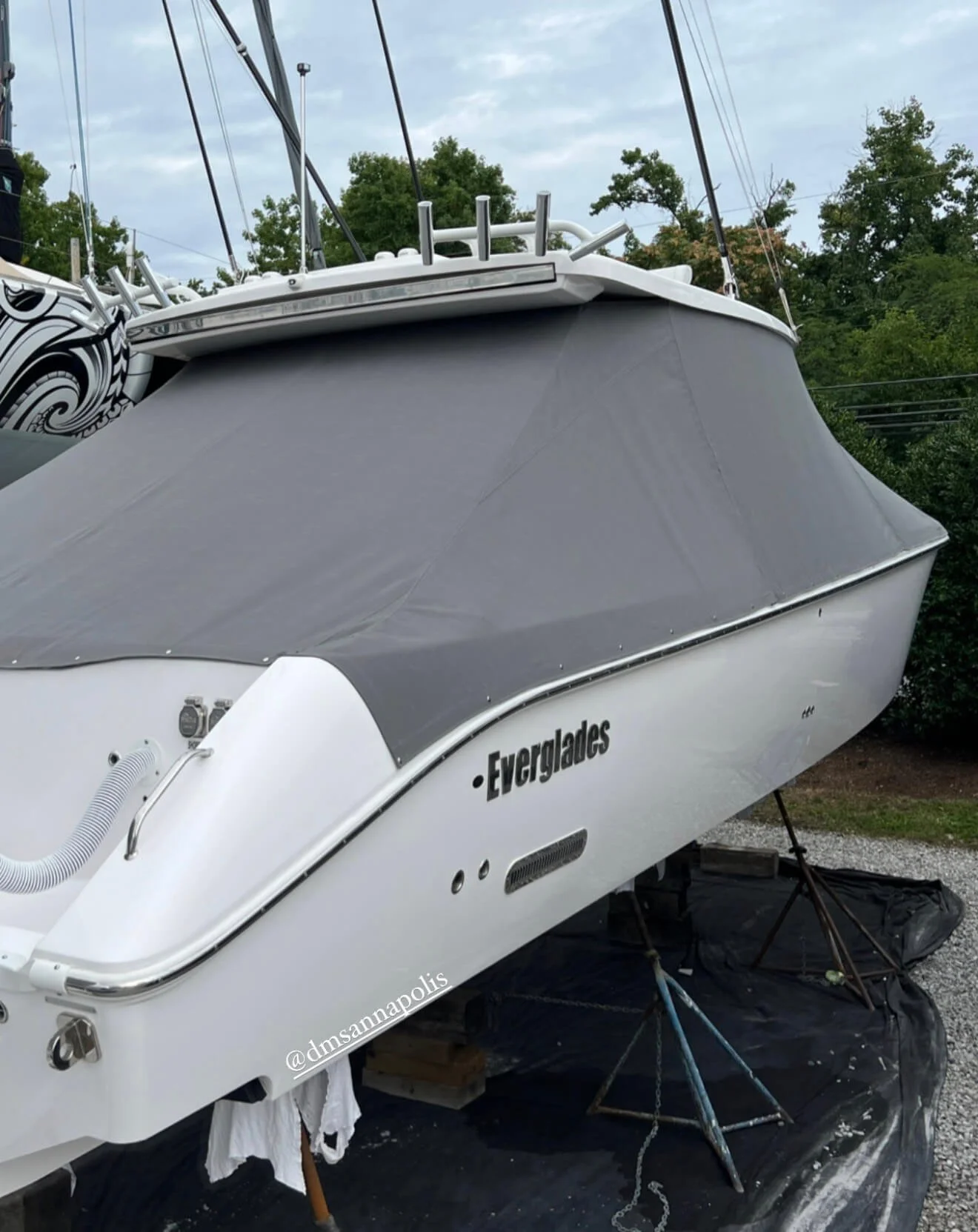 A white boat named Evergreen on land with a gray cover over its deck, supported by metal stands on a black tarp, with trees and a cloudy sky in the background.