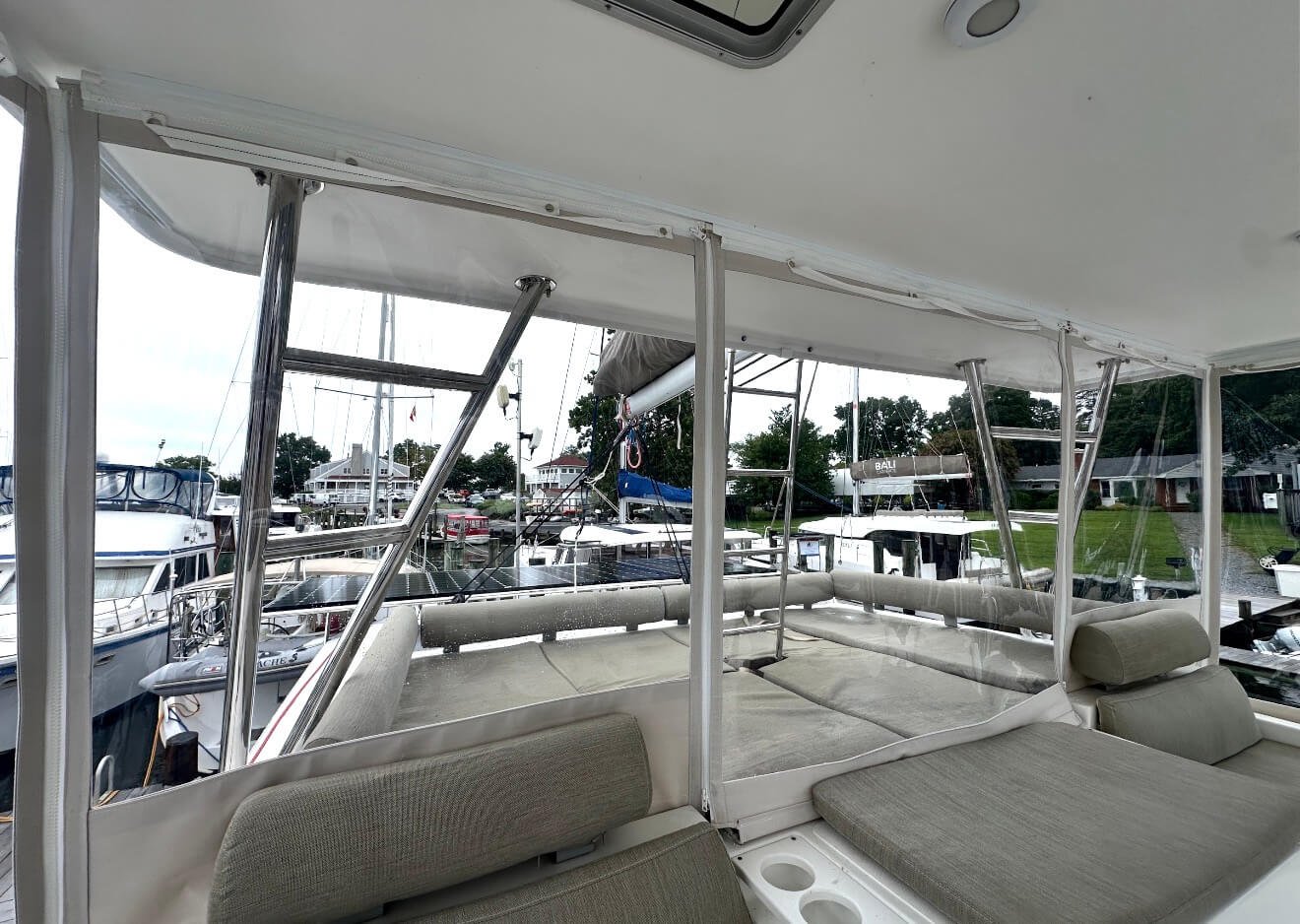 View from inside a boat's covered deck area showing a marina with anchored boats and sailboats, with green grass and trees in the background.