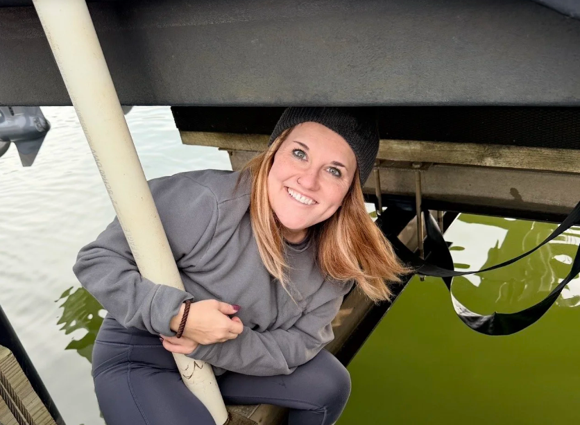 Woman with long red hair, wearing a black beanie and gray hoodie, smiling while on a boat near the water, holding onto a white pole with water below.