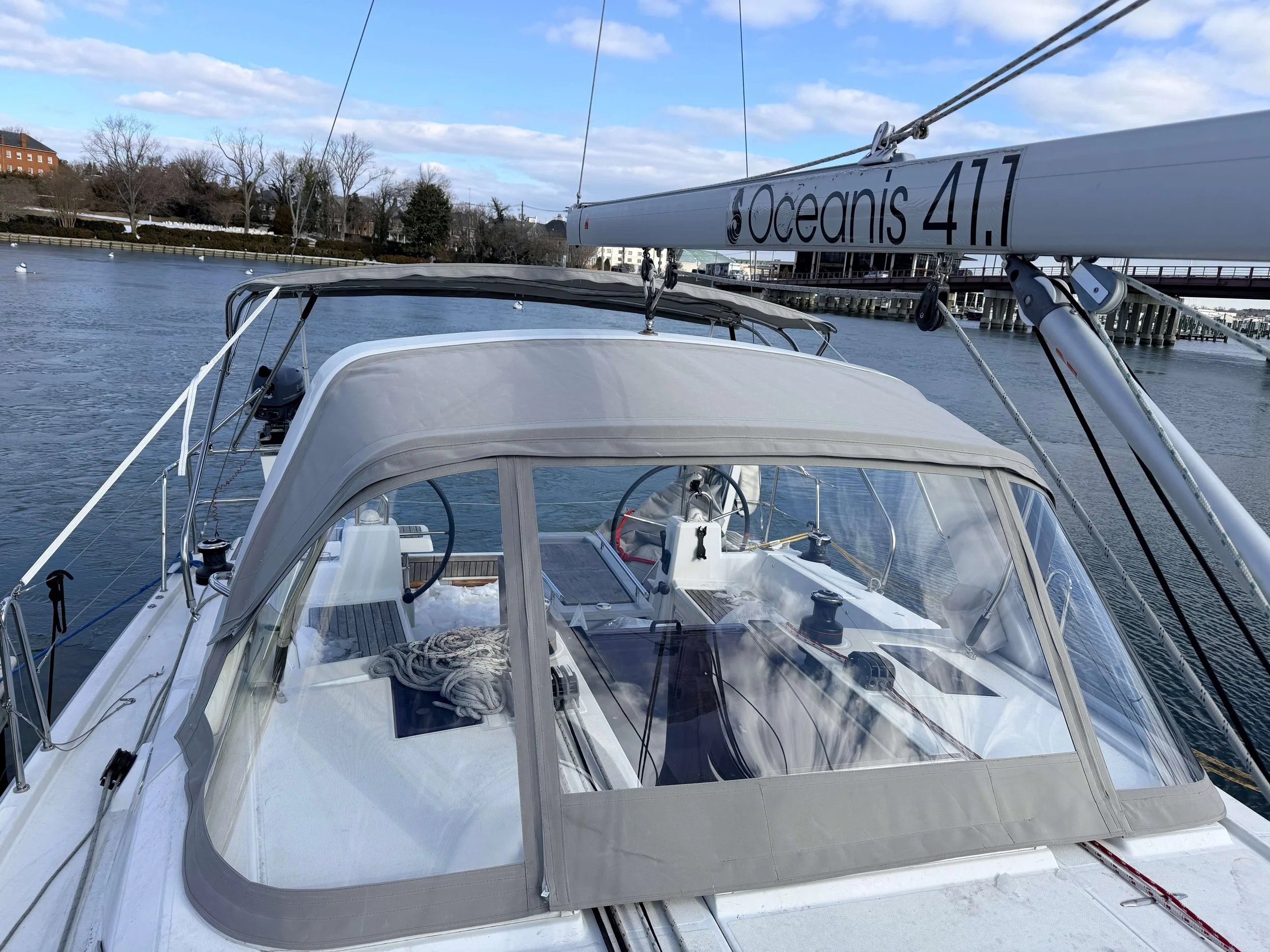 View of a sailboat with a cabin and rigging, sailing on a calm waterway under a partly cloudy sky. Trees and a bridge are visible in the background.