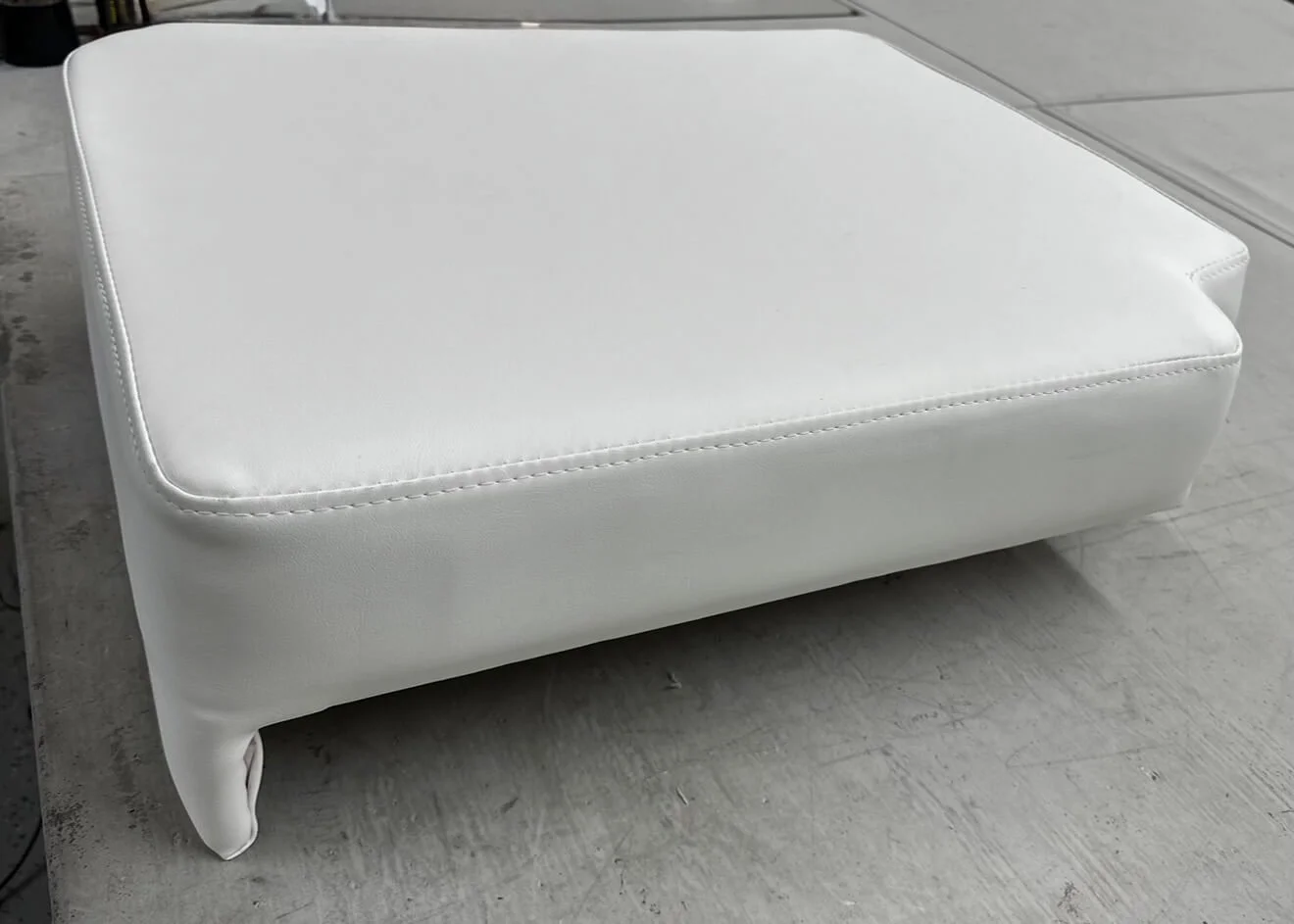 A white upholstered ottoman with rounded edges and curved wooden legs, placed on a concrete floor.