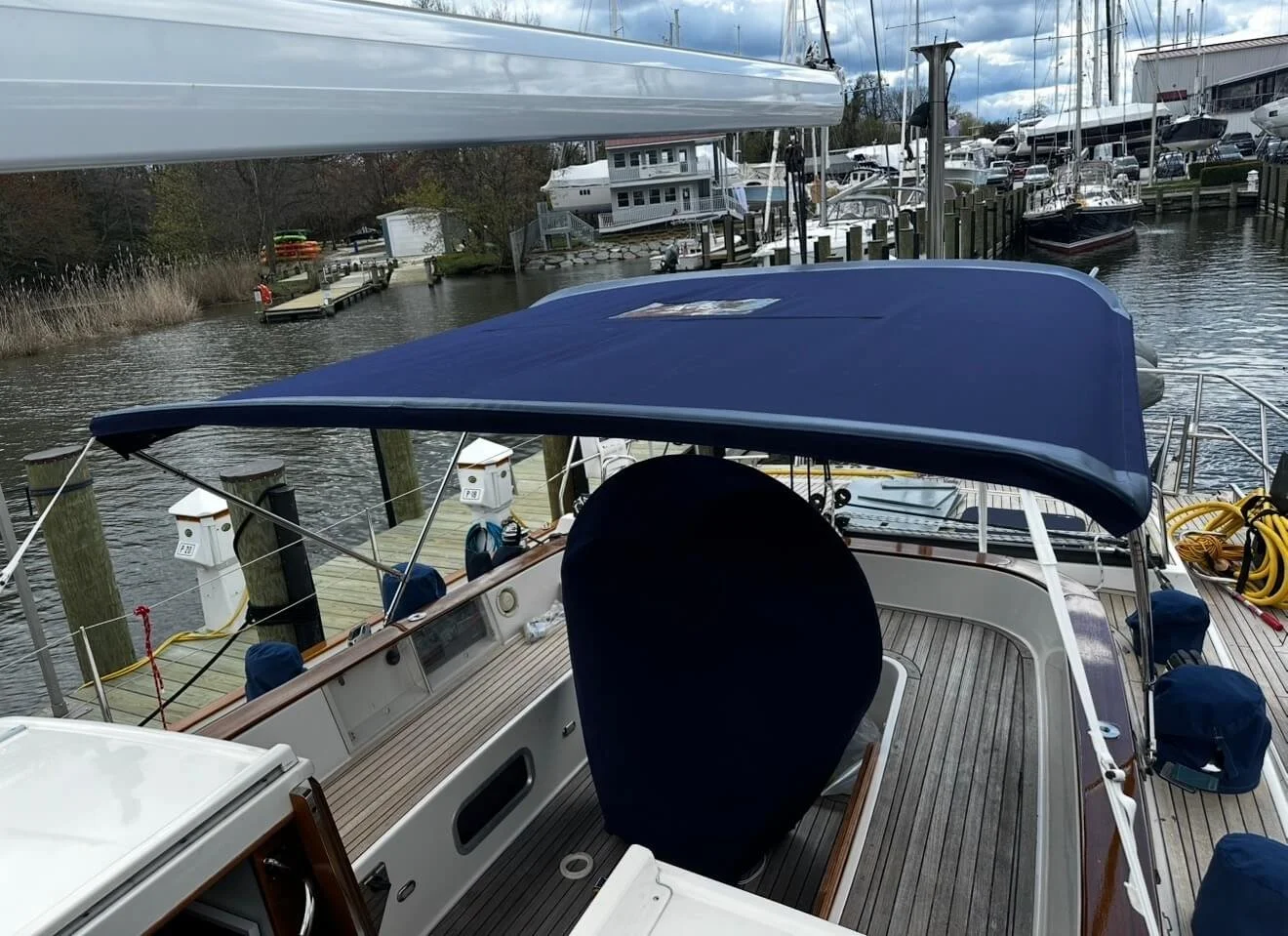 View of a marina with boats docked, including a yacht with a blue canopy, wooden dock, and other sailboats in the background.