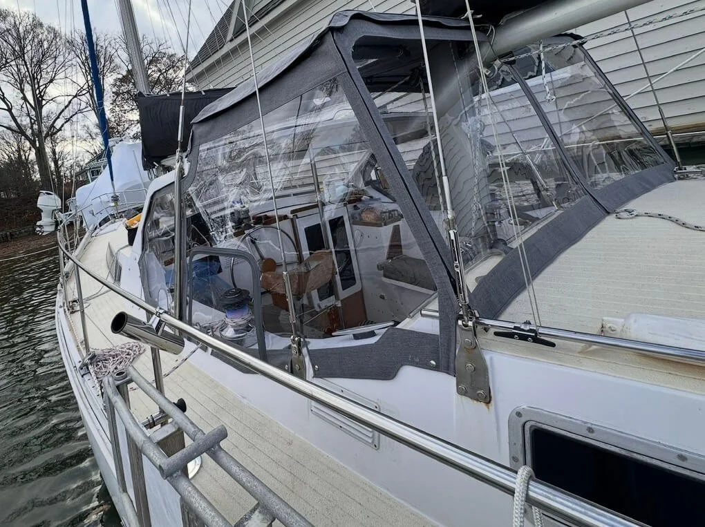 A sailboat docked at a marina with a gray canvas cover over the cockpit and clear plastic windows, surrounded by water and trees in the background.