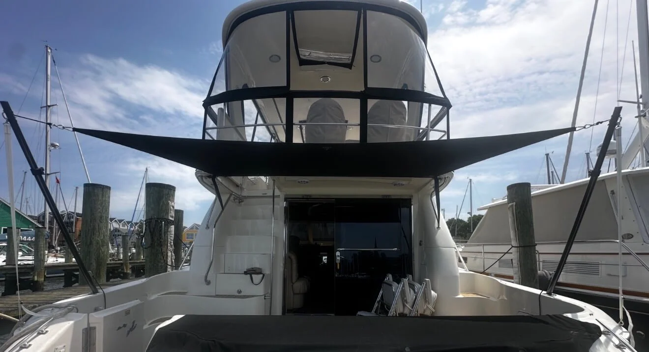Front view of a white yacht docked at a marina, with a boat's upper deck and a black canopy, surrounded by other boats and wooden pilings.