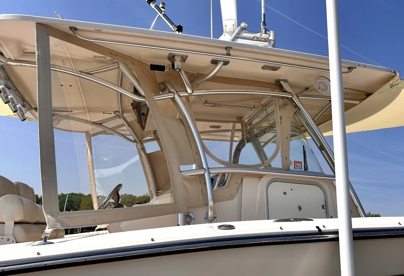Close-up view of a boat's control area with steering wheel, metal bars, and canopy, against a background of clear blue sky and distant trees.