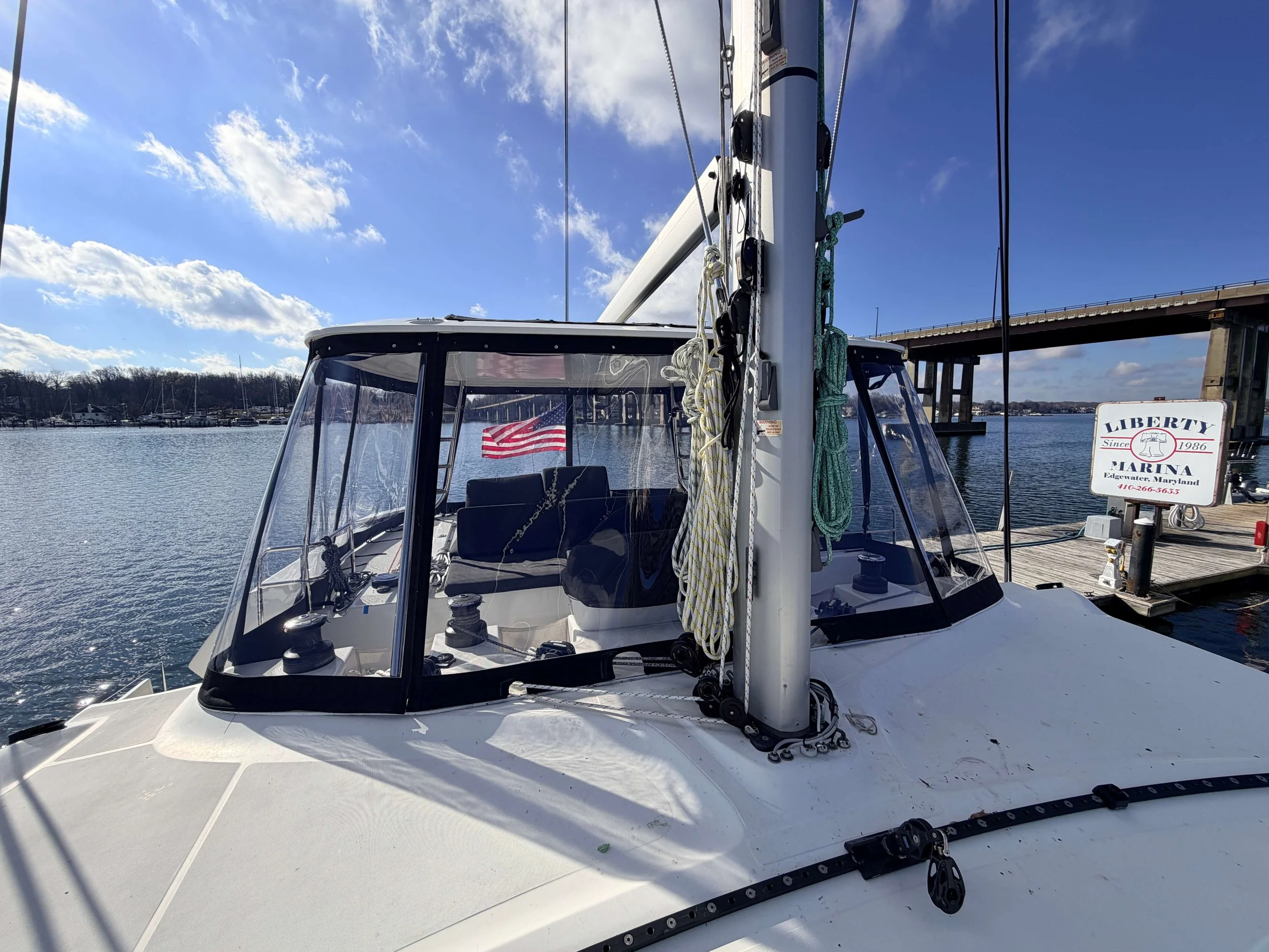 View of a sailboat docked at a marina with an American flag visible through the window, a bridge in the background, and a sign reading "Liberty Marina".