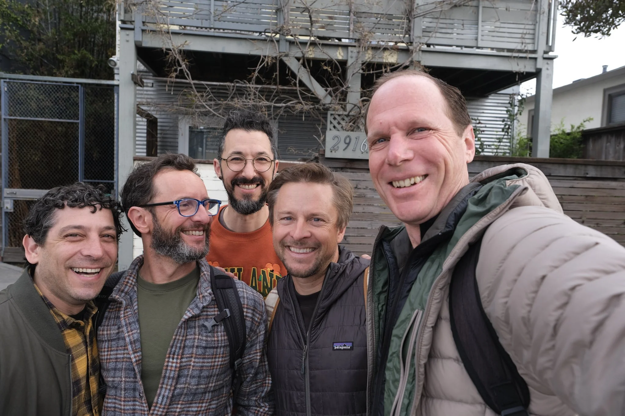 Five smiling men taking a group selfie outdoors in front of a house with a wooden and metal exterior and leafless branches. They are wearing casual outdoor jackets and carrying backpacks.