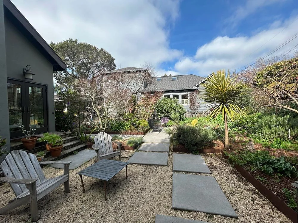 A backyard garden with a concrete pathway, wooden benches, potted plants, trees, and lush greenery under a partly cloudy sky.