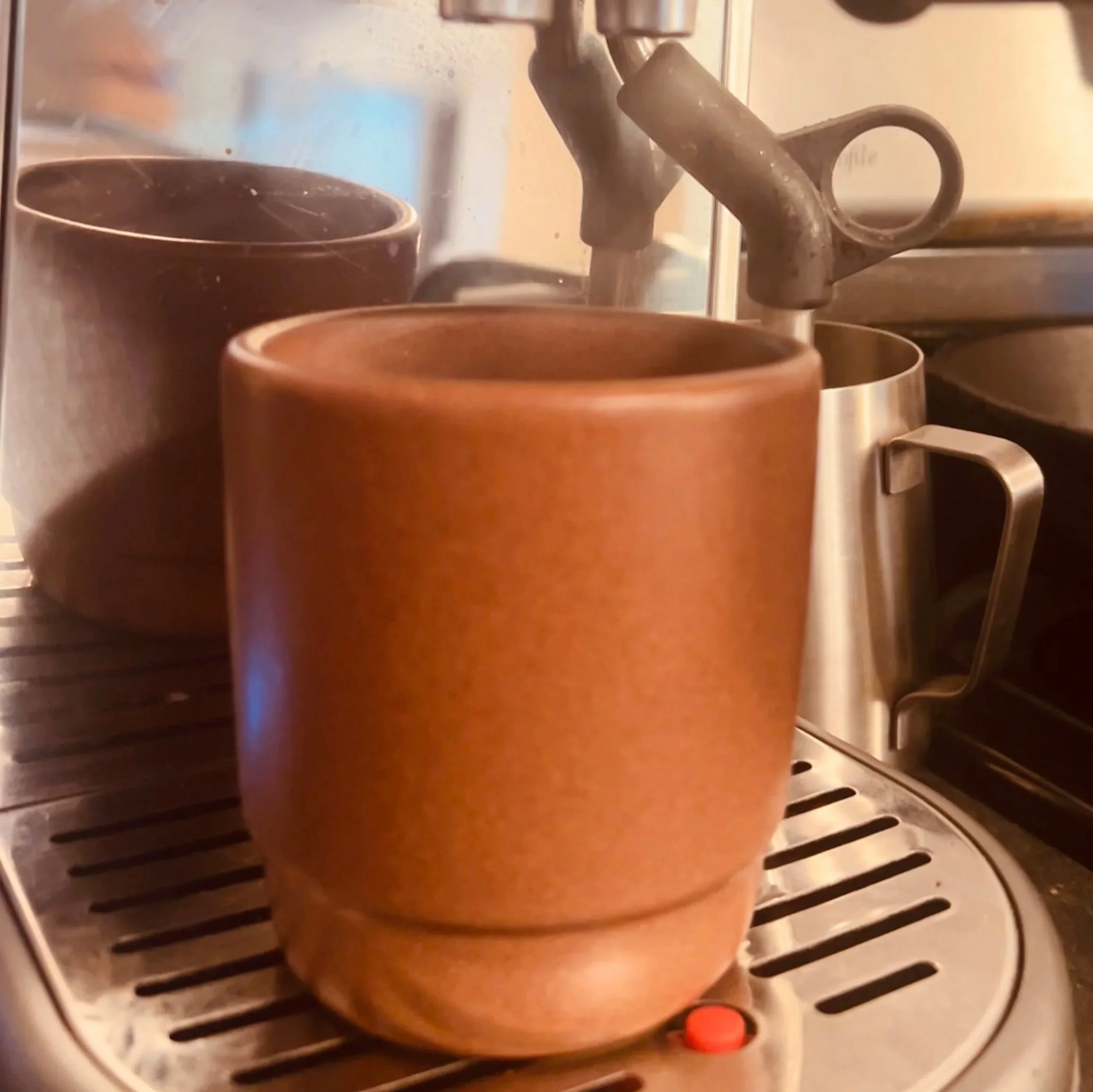 A brown ceramic mug placed on the drip tray of an espresso machine, with a metal milk frothing pitcher and another cup in the background.