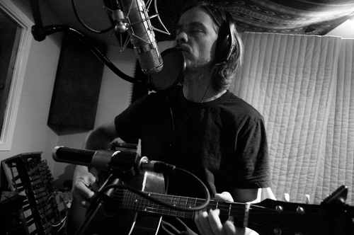 A man singing into a microphone while playing an acoustic guitar in a recording studio.