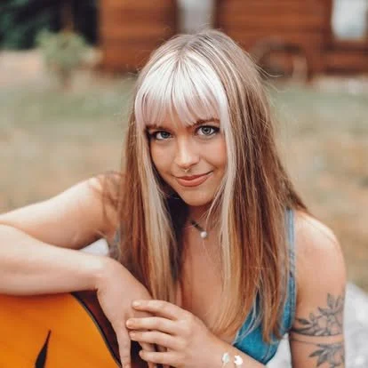 A young woman with long blonde hair and bangs, holding a guitar, outdoors with a blurred background.