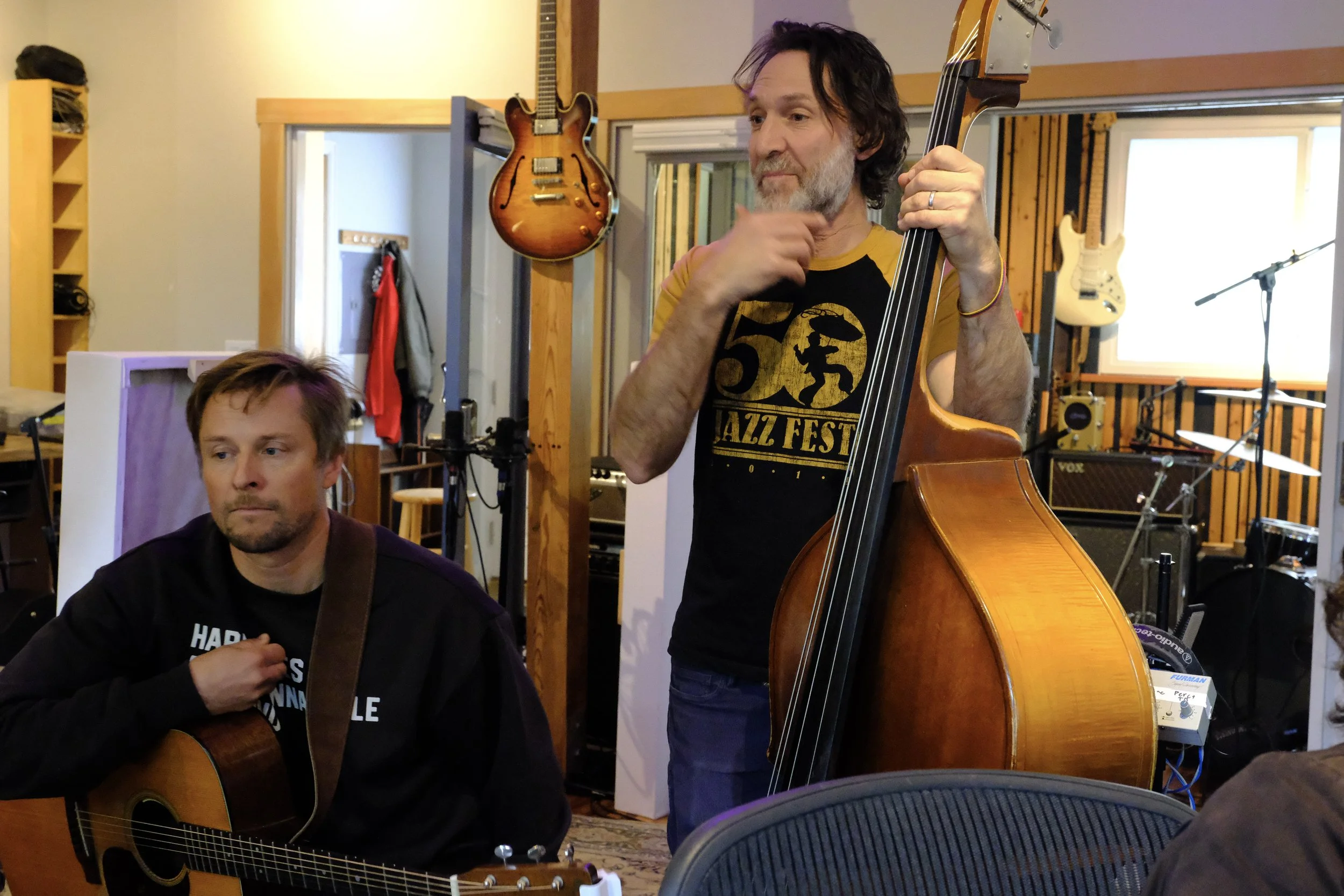 Two musicians in a recording studio, one playing an acoustic guitar and the other playing a double bass, surrounded by musical equipment and guitars hanging on the wall.