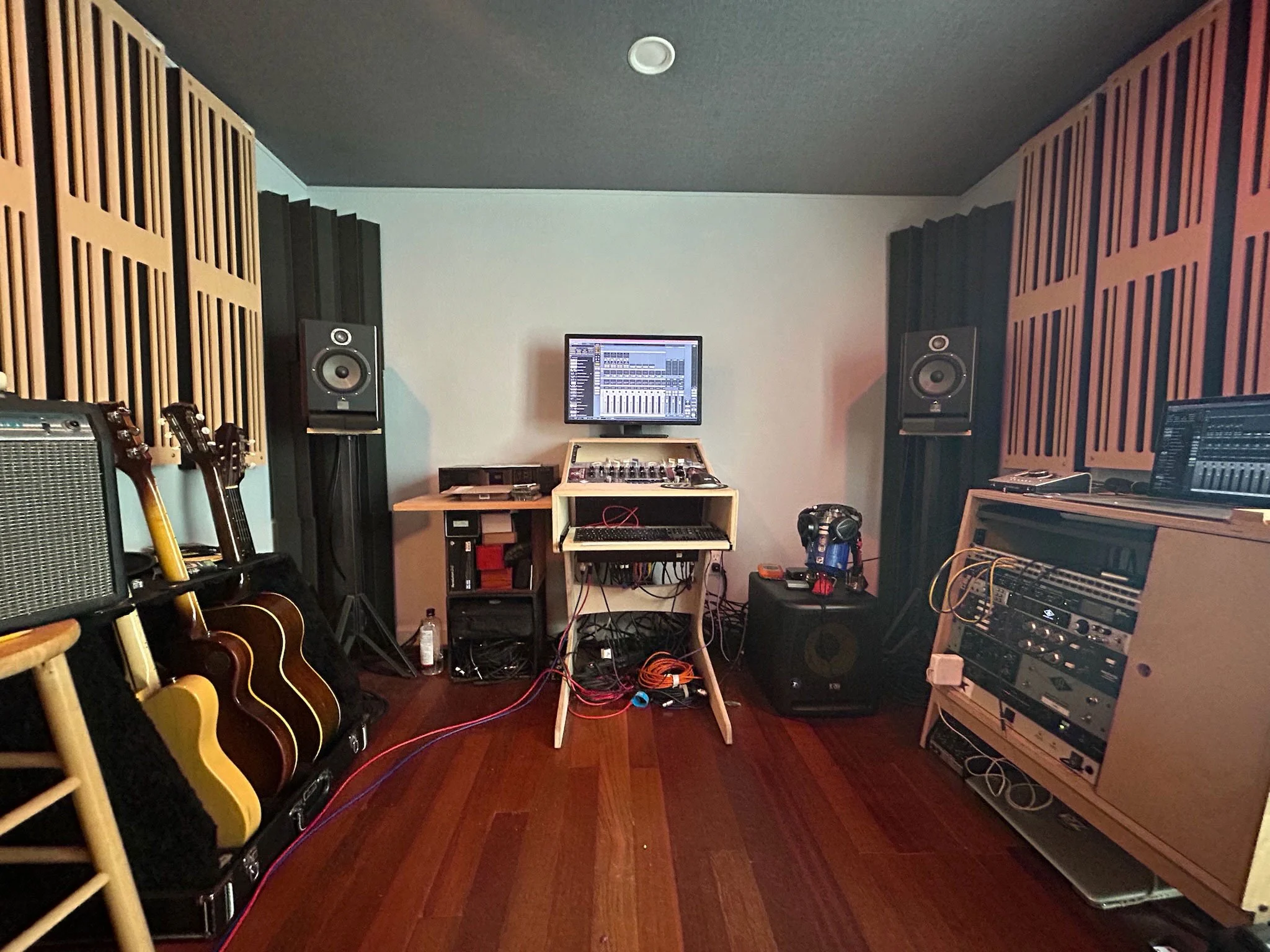 Inside Ben's music studio with guitars on the left, acoustic foam panels on the walls, a computer monitor displaying digital audio workstation on a table, and audio equipment including speakers, mixer, and audio interfaces.