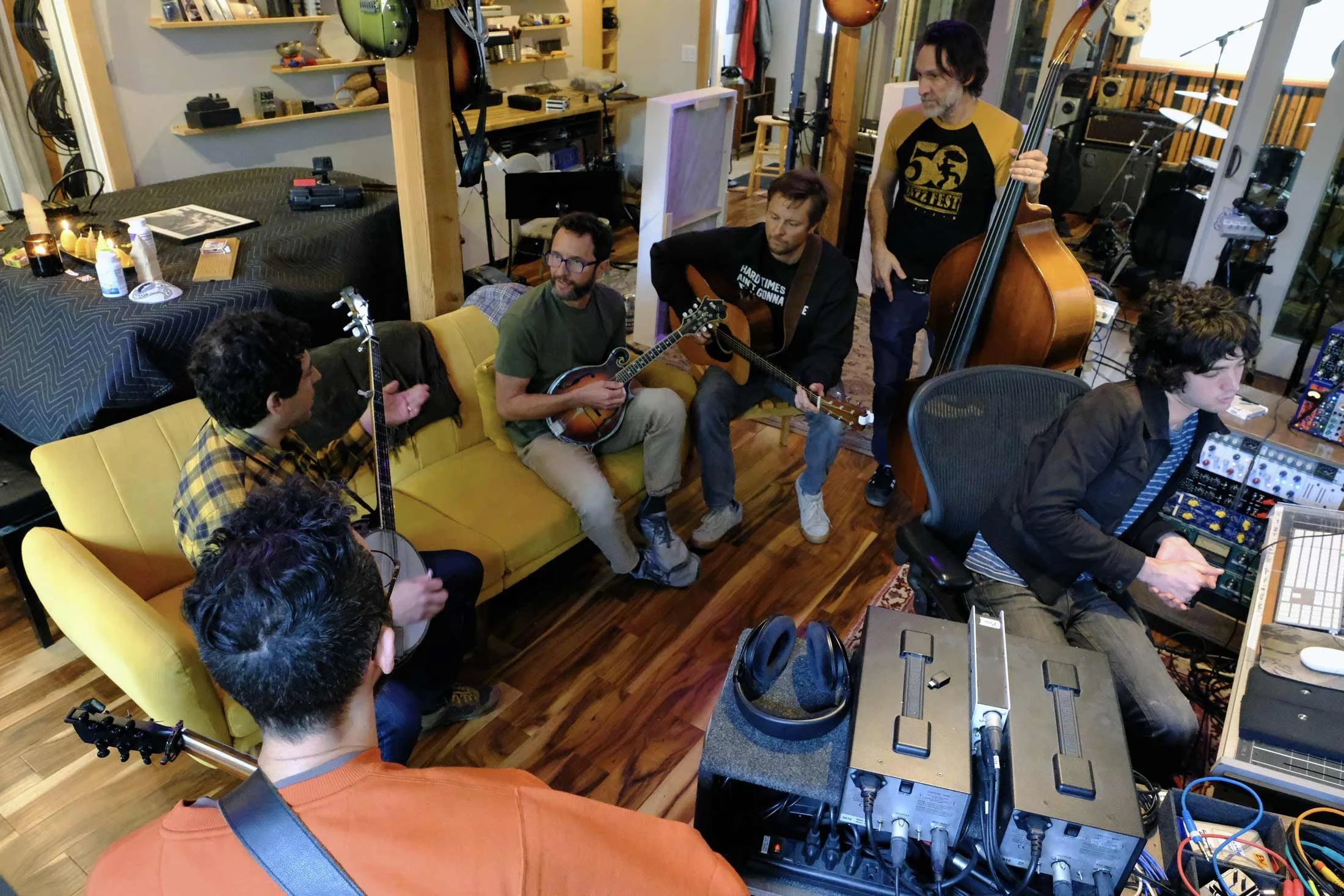 A group of five musicians in a recording studio. Two are playing guitars, one is playing an upright bass, and one is working with electronic music equipment. The room has wooden floors and shelves with various instruments and equipment.