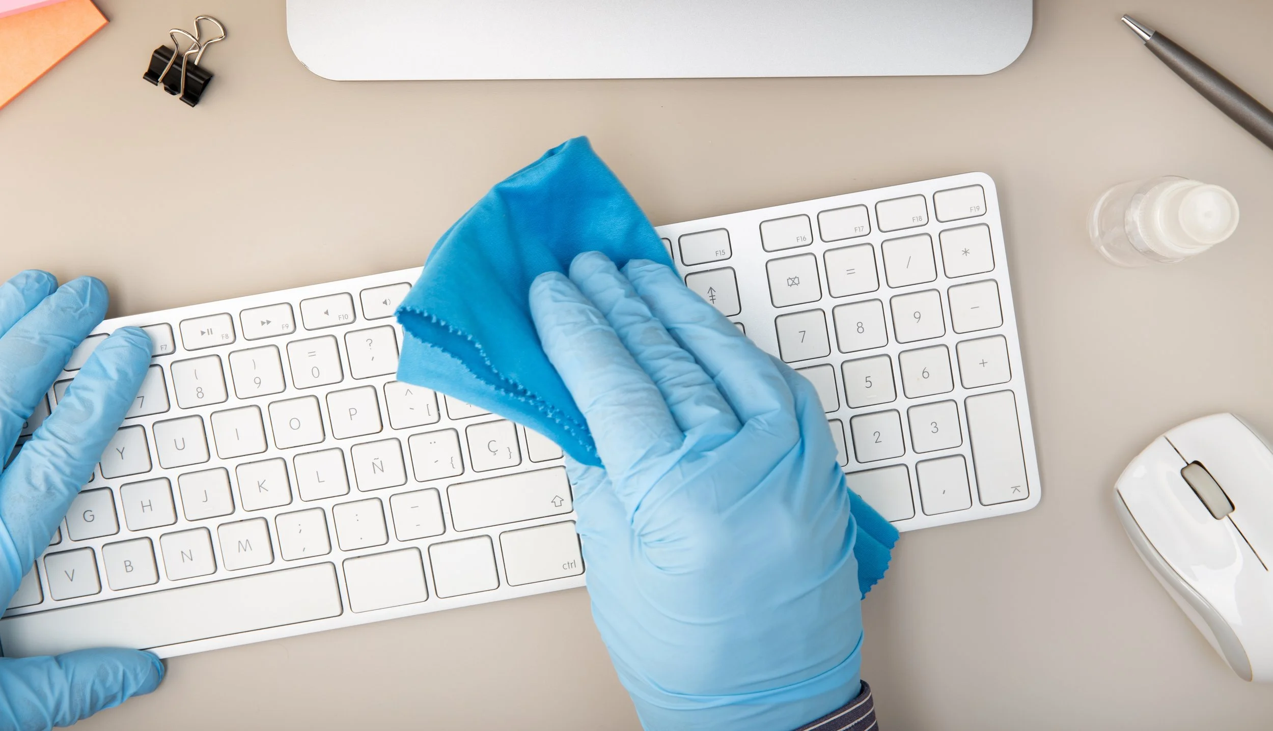Person wearing blue gloves cleaning a white computer keyboard with a blue cloth on a desk.
