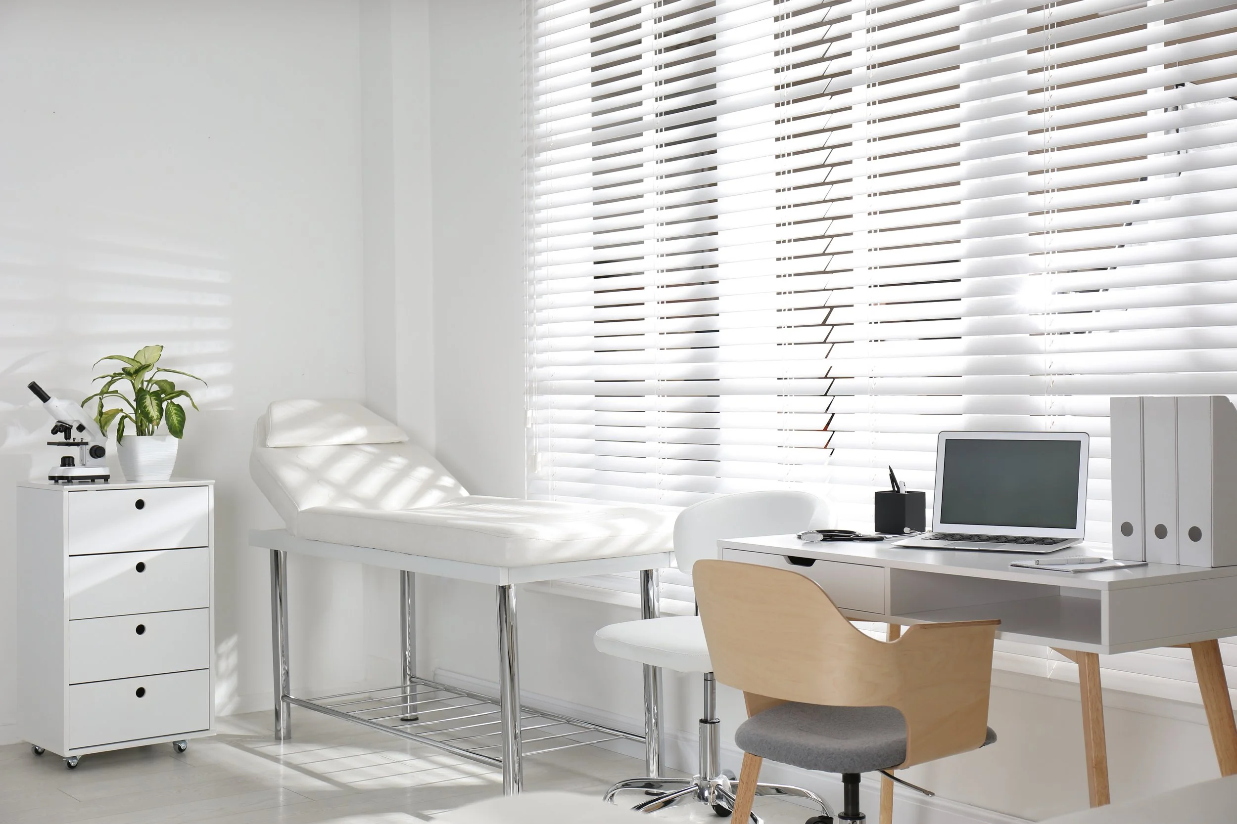 A medical examination room with a white examination table, a white desk with a laptop, office supplies, and a computer, a wooden chair, and a small white drawer with a potted plant and microscope. Bright sunlight coming through white blinds.