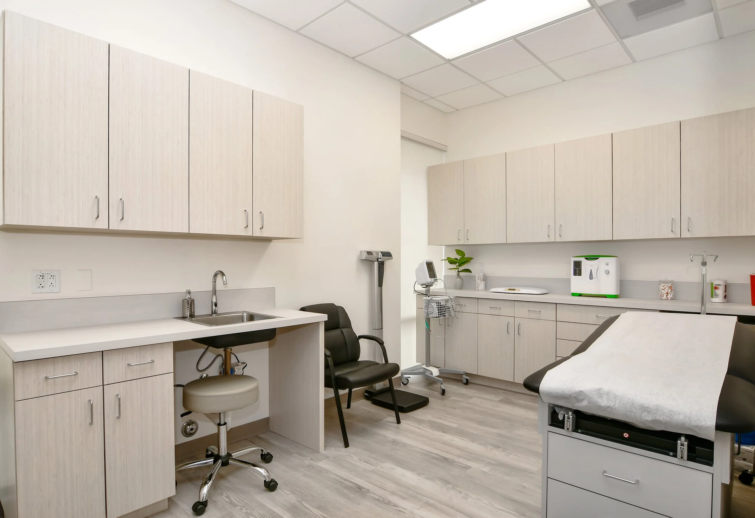 Medical examination room with cabinets, countertops, a sink, medical equipment, and examination bed.