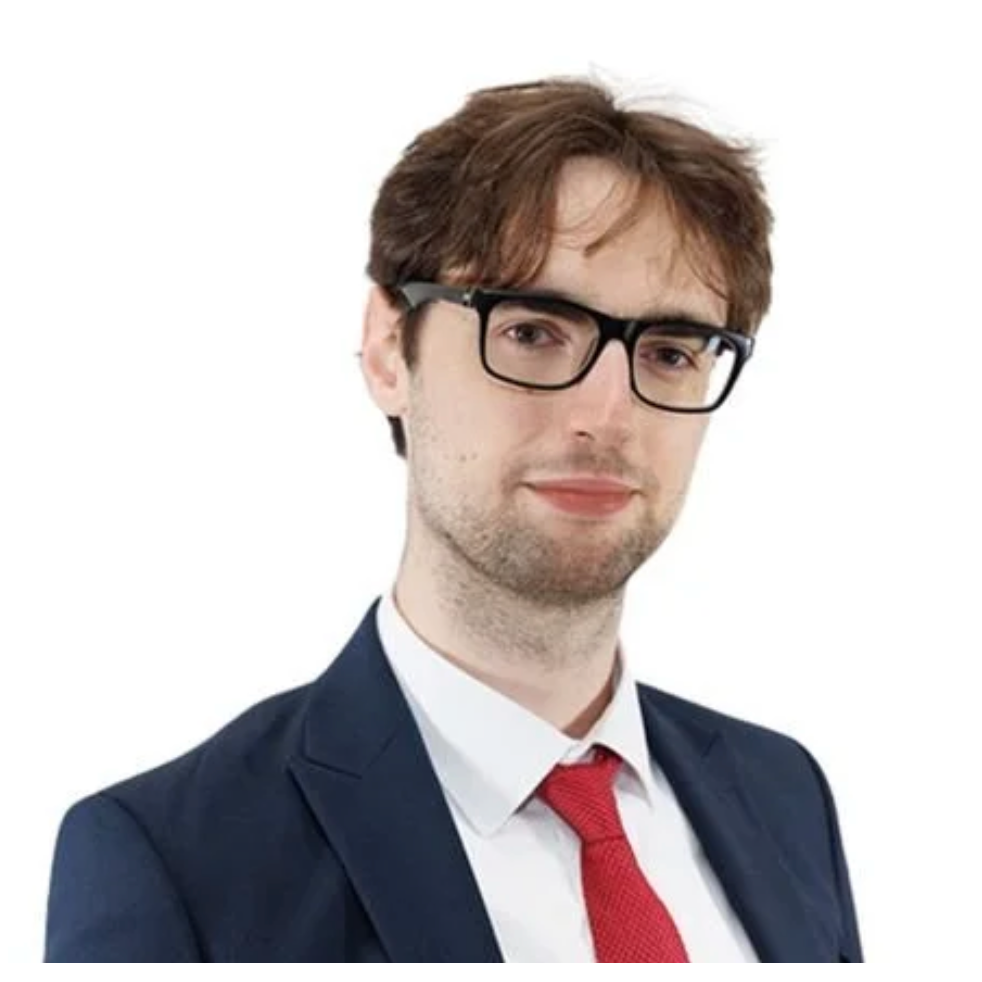 A young man with brown hair wearing black glasses, a navy suit, white shirt, and red tie, against a plain white background.
