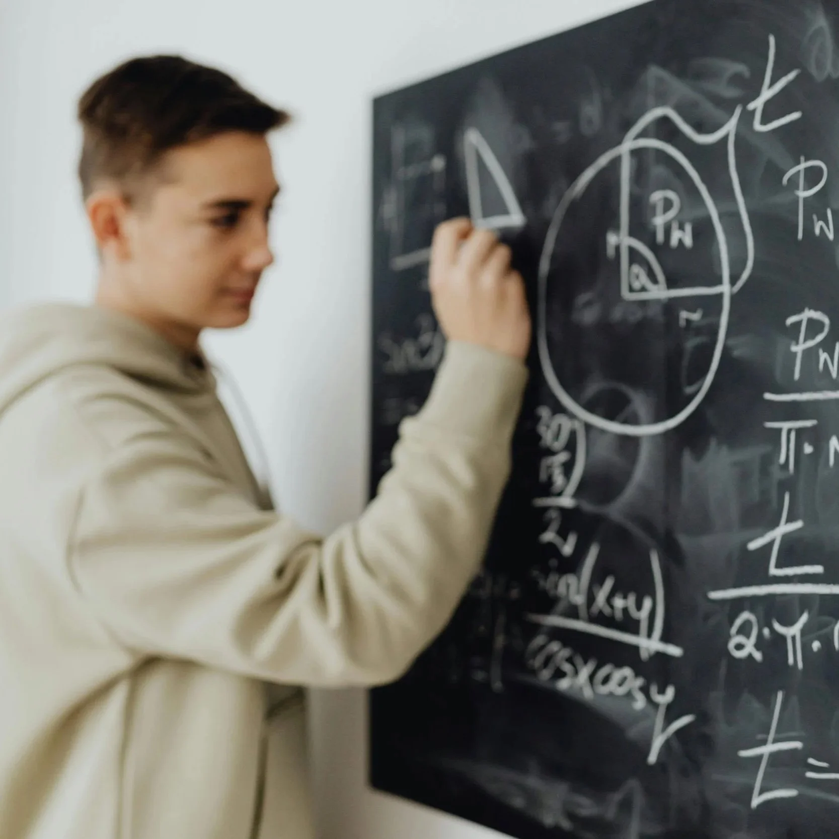 A young man writing mathematical equations and diagrams on a black chalkboard.