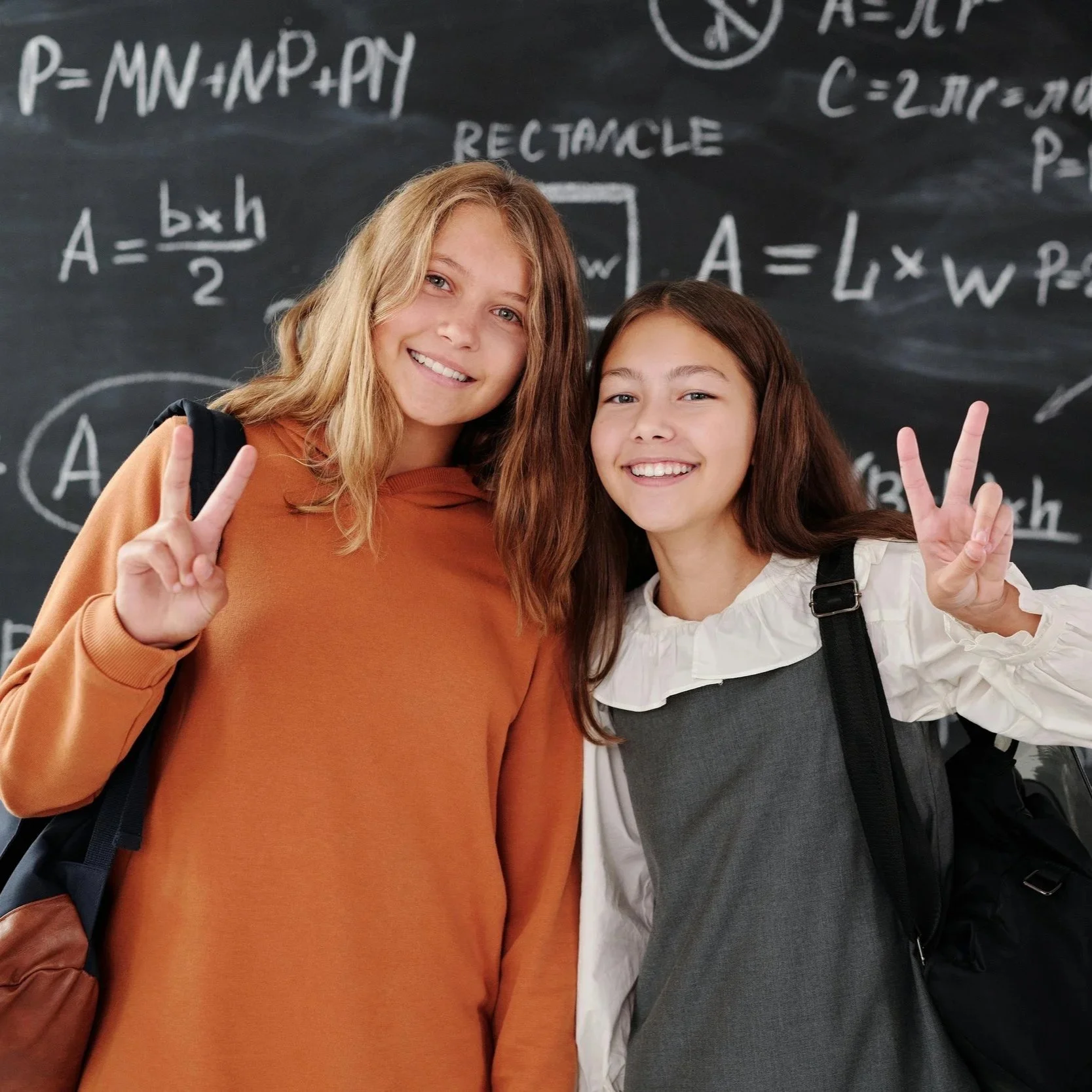 Two smiling girls making peace signs while standing in front of a blackboard filled with mathematical and scientific equations.