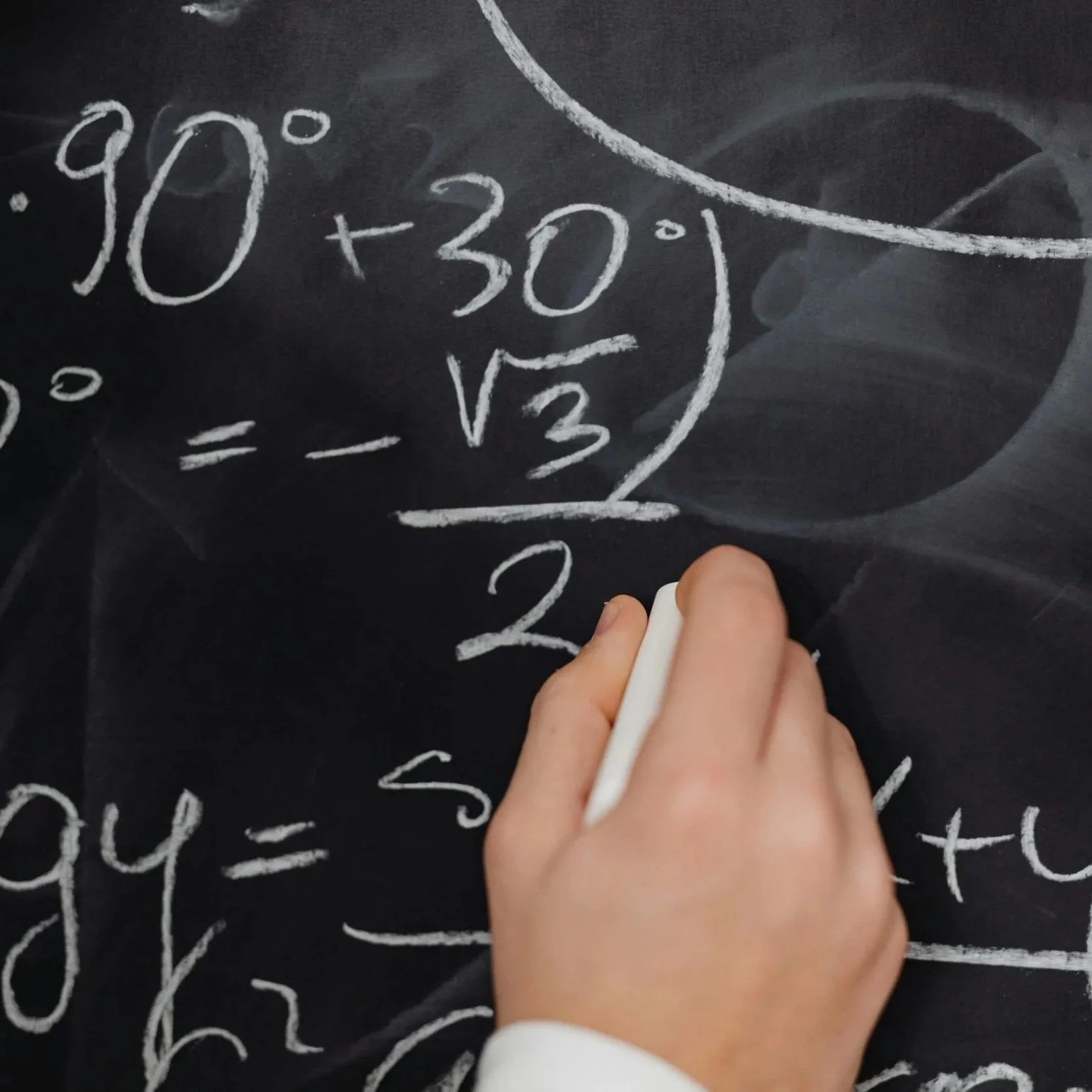 Close-up of a hand writing elliptical mathematical expressions on a black chalkboard with white chalk, featuring fractions, square roots, plus and equal signs.
