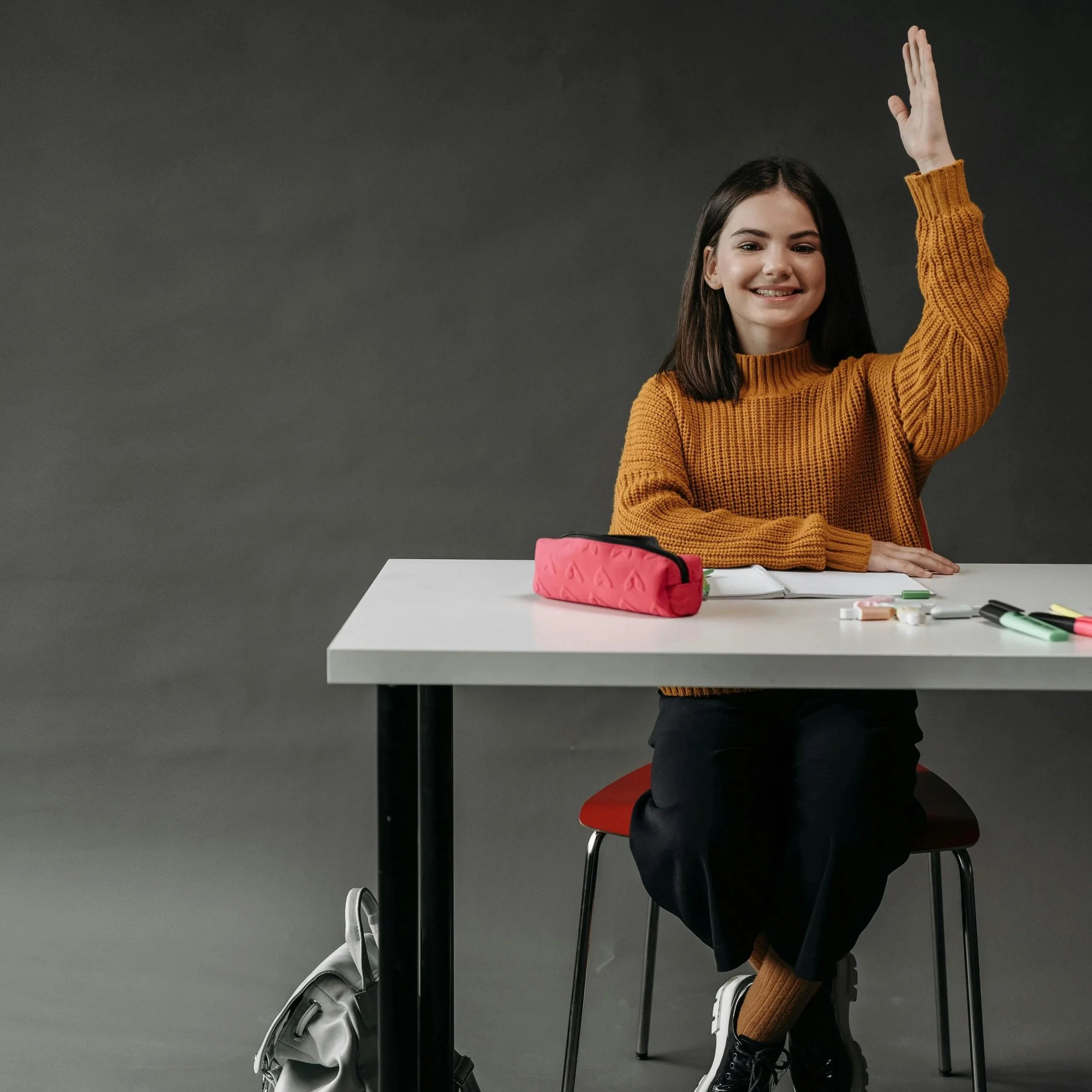 Young woman with dark hair wearing a mustard-colored sweater, sitting at a classroom desk with a pink pencil case, notebook, and pens, raising her hand, and smiling. There is a backpack on the floor under the desk and a gray backpack leaning against the side of the desk.