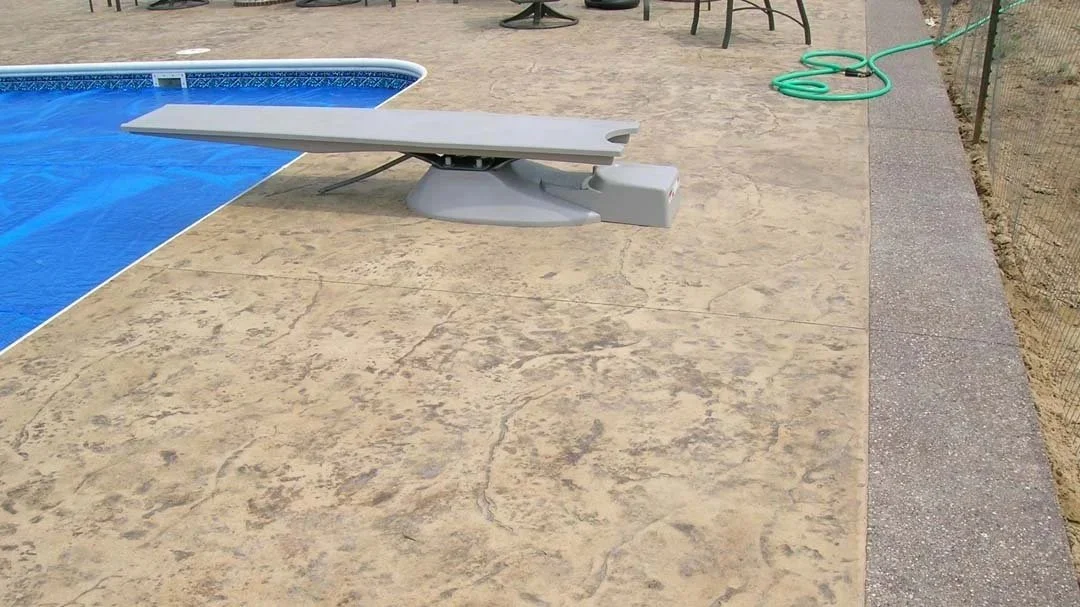 Concrete pool deck with a diving board, green garden hose, and outdoor chairs in the background.