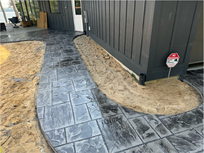 Recently installed gray stamped concrete pathway around the building with a small sand area and a 'No Trespassing' sign.