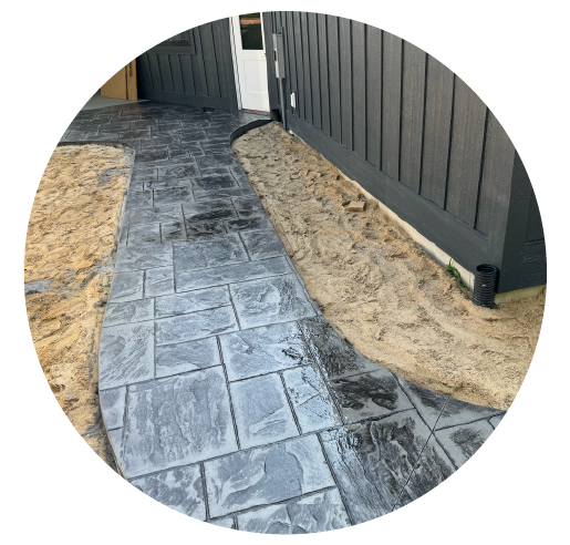 Newly installed stamped concrete walkway with a wood grain pattern next to a gray fence and a house entrance, with sand on the sides awaiting finishing.