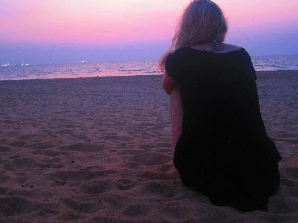 A woman with long hair sitting on a sandy beach at sunset, facing the ocean with pink and purple hues in the sky.