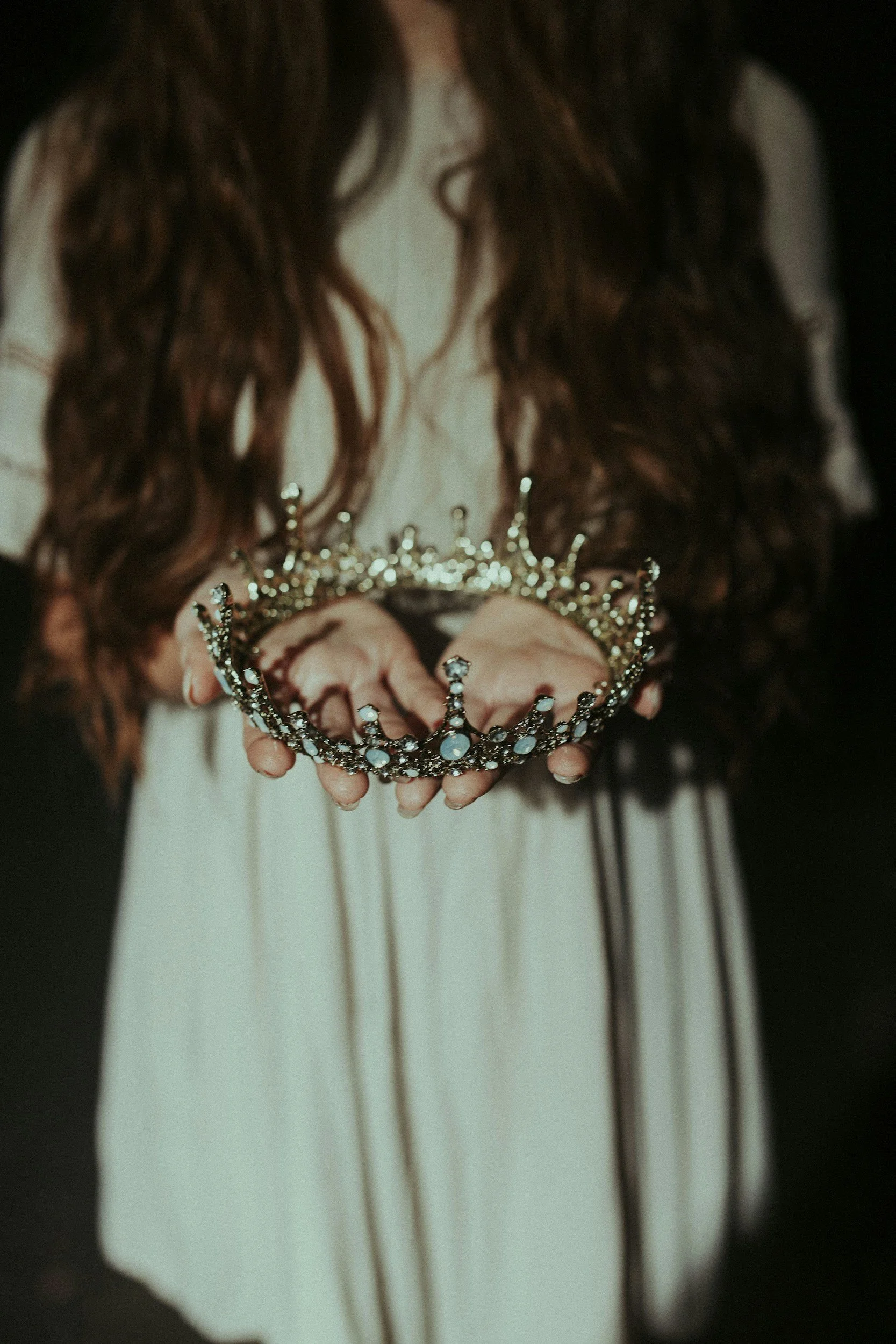 A person with long brown hair in a white dress holding a silver tiara with blue stones against a dark background. The Wild Rose Chronicles Membership | Celestial Women's Studies Classes