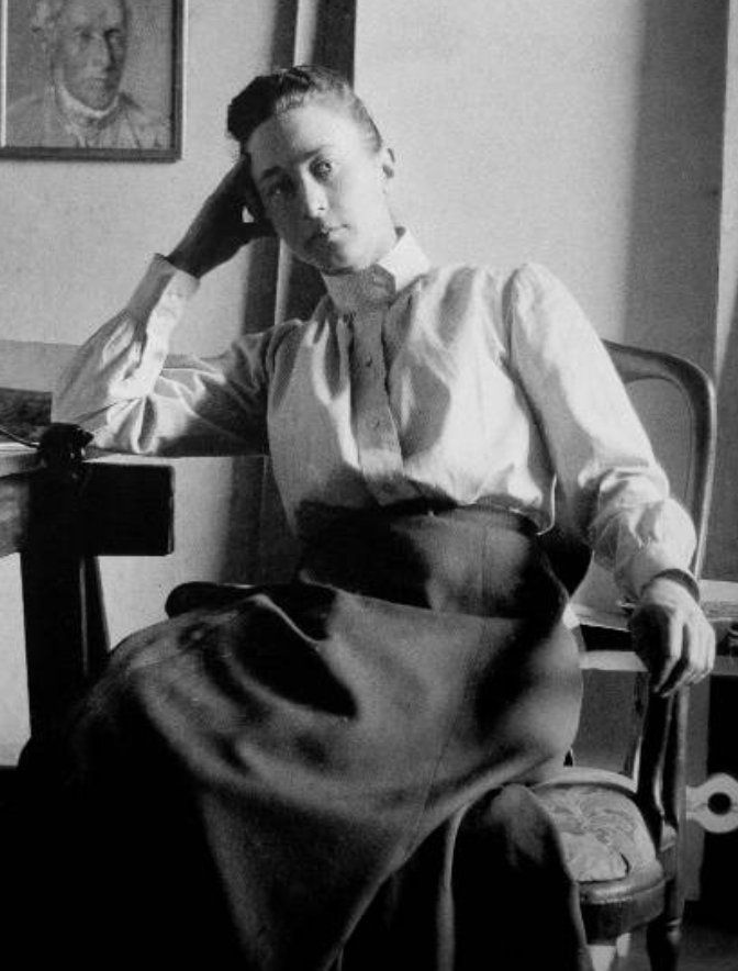 A black-and-white photo of Hilma Af Klint sitting on a chair, resting her head on her hand, wearing a blouse and a skirt, with a portrait hanging on the wall behind her. The Wild Rose Chronicles Membership | Celestial Women's Studies Classes
