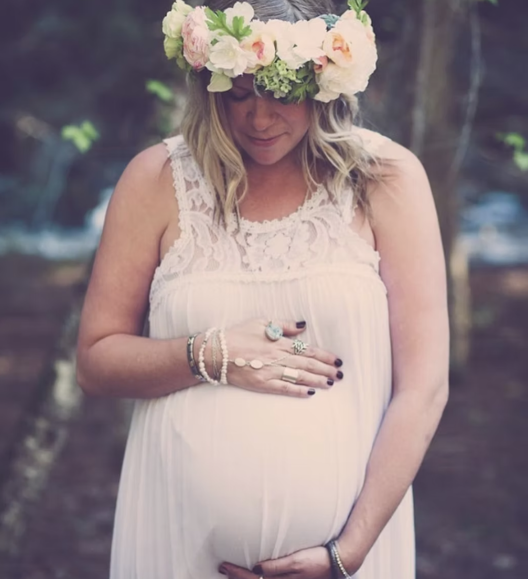 Pregnant woman wearing a flower crown and white lace dress, holding her belly with one hand and resting her other hand on her stomach, outdoors.