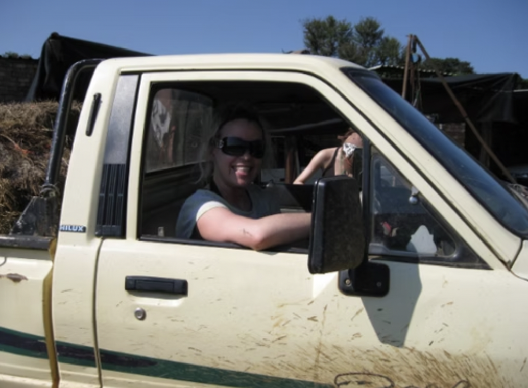A woman with blonde hair, wearing sunglasses and a gray shirt, smiling and sitting in the driver's seat of a small, off-white pickup truck with some dirt or scratches on the door. Another person wearing a mask and a tank top is visible in the backgro