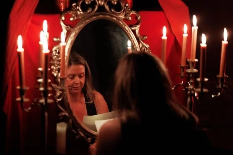 A woman with long hair reads a letter or paper in front of a mirror, surrounded by lit candles and red drapes, creating an intimate, romantic atmosphere for the Inner Union journey. Inner Union Online Course.