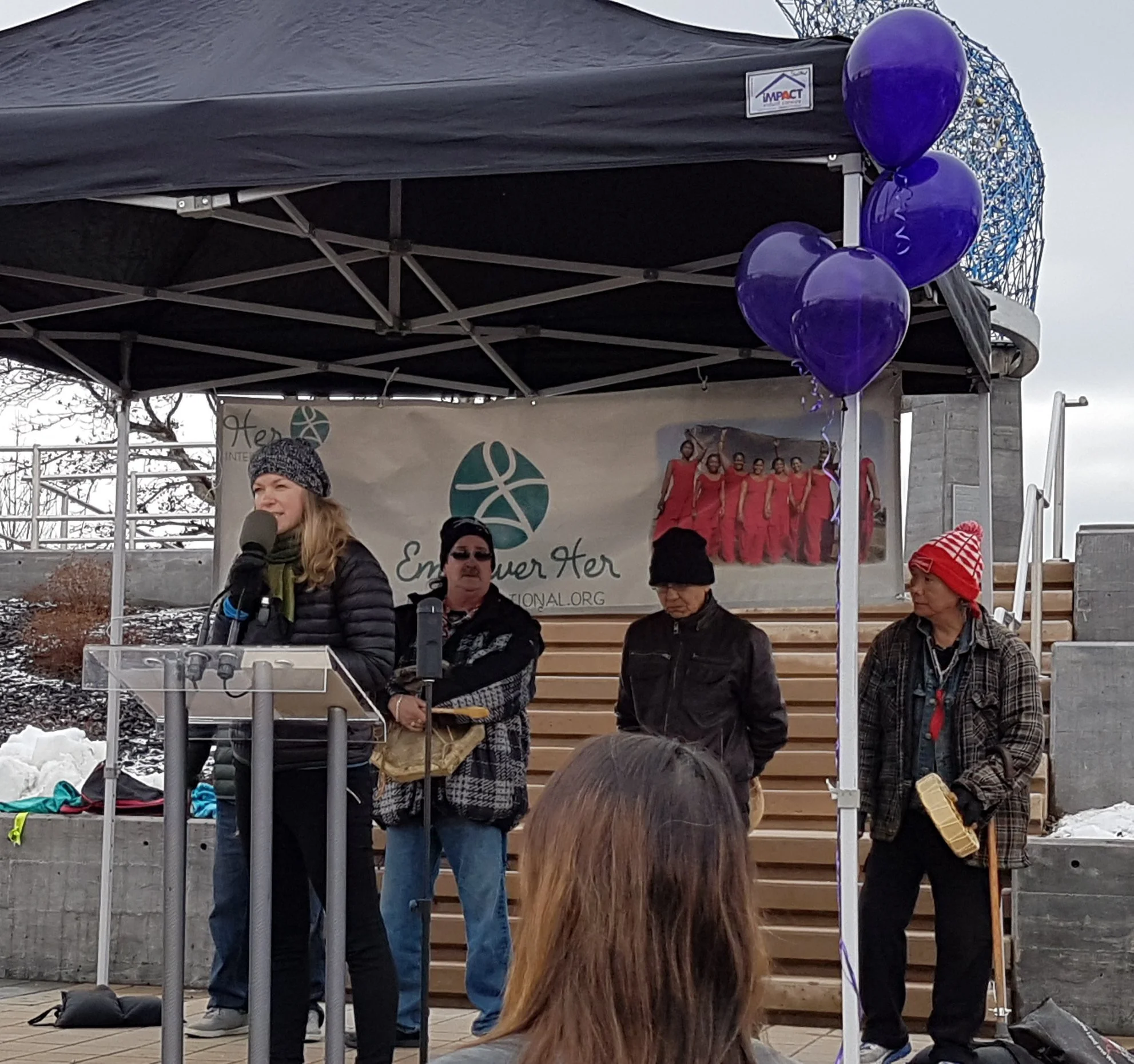 People standing on a stage outdoors, some wearing winter clothing, with a banner and purple balloons. The sky is overcast, and there are trees without leaves in the background.