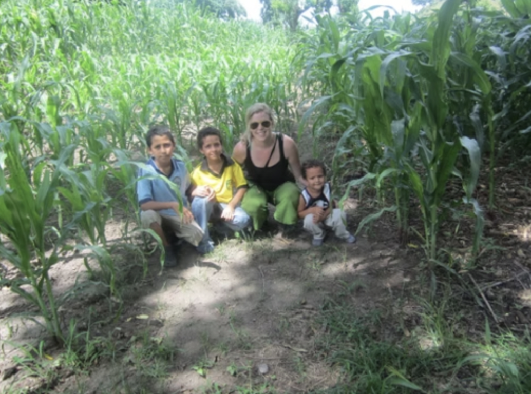 A woman and three young boys kneeling in a field of tall green corn plants.
