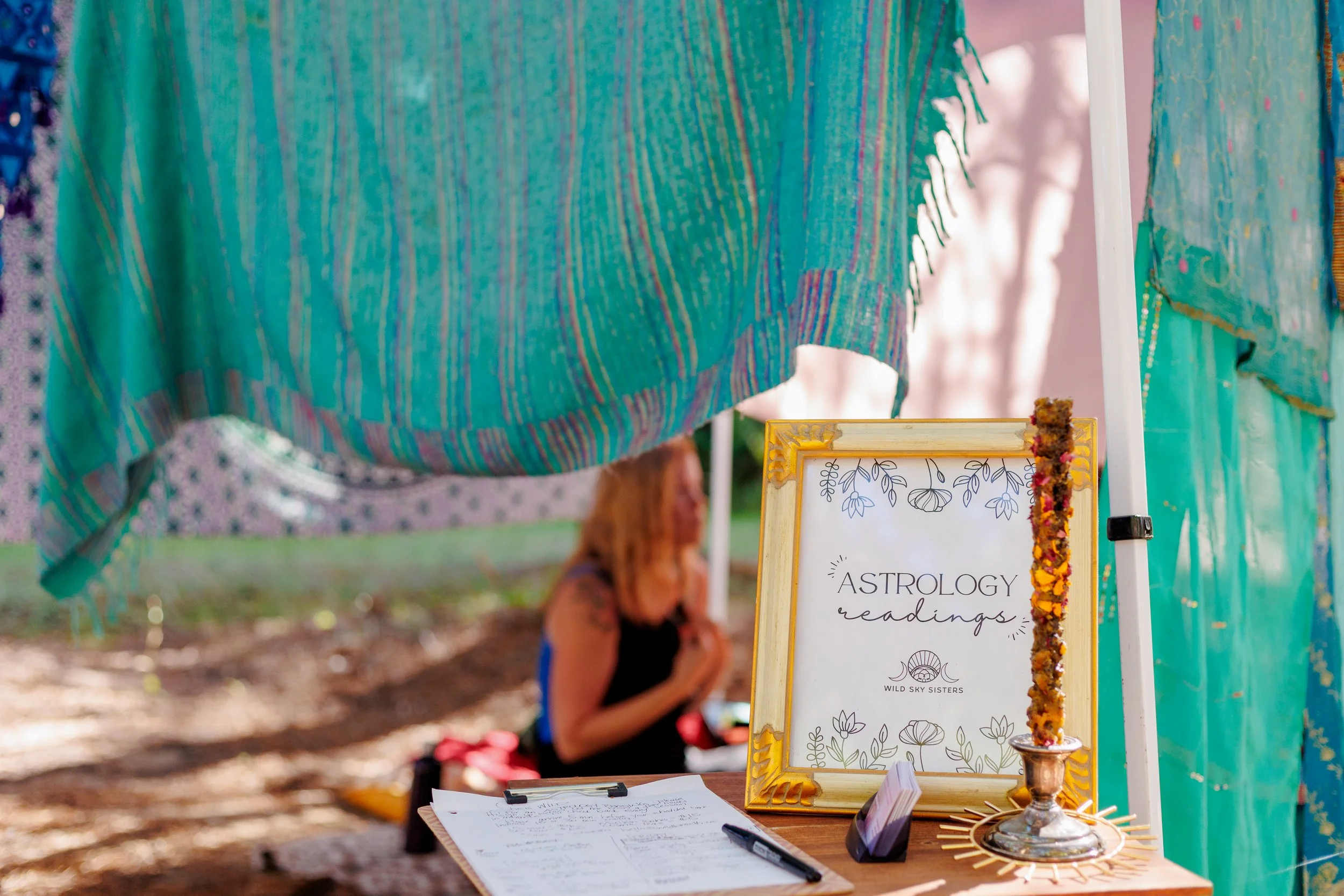 A table at an outdoor astrology readings booth with a framed sign, a chalice, papers, a pen, and a decorative garland. A person sits in the background with a colorful scarf. Brightly colored tents and trees are visible.