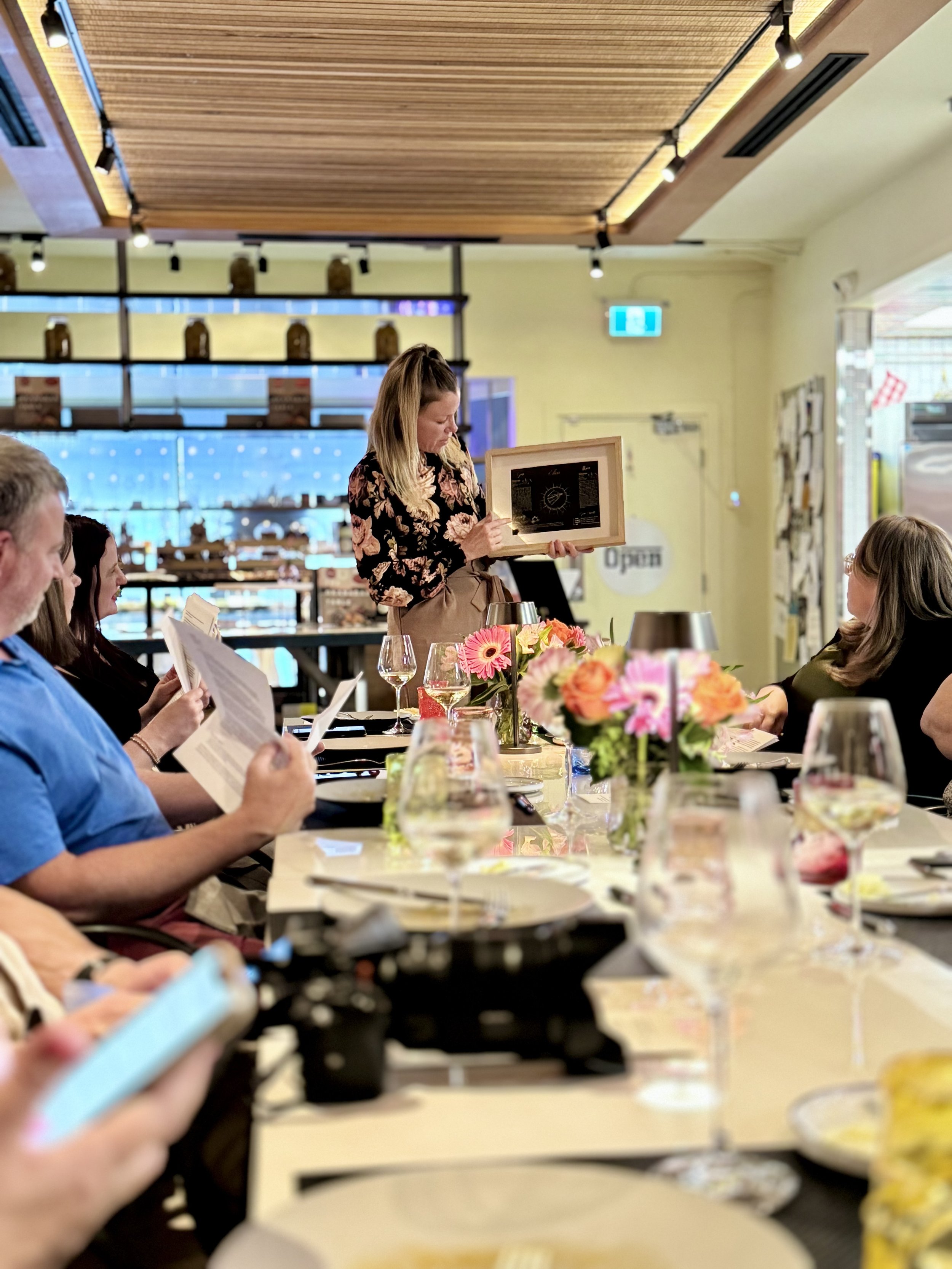 A woman in a black floral shirt stands at a table holding a framed menu or piece of artwork, while seated guests read menus at a dinner or event, with flower arrangements and wine glasses on the table.