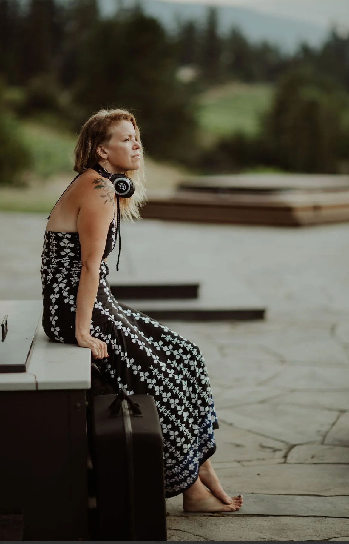 Tamara McLellan wearing a dress, sitting barefoot on a bench outside, with a black tattoo on her left shoulder, listening to music with headphones around her neck. She looks contemplative, with a scenic green and cloudy background.