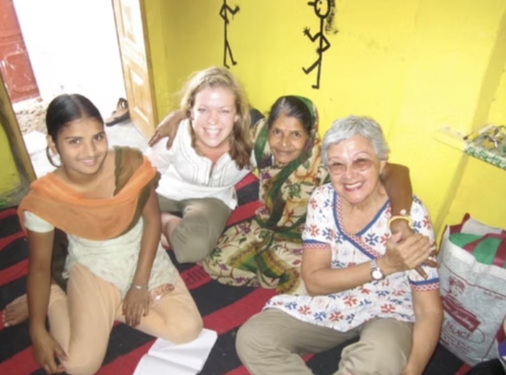 Four women of different ages sitting on a colorful mat in a room with yellow walls, smiling, with two stick figure drawings on the wall behind them.