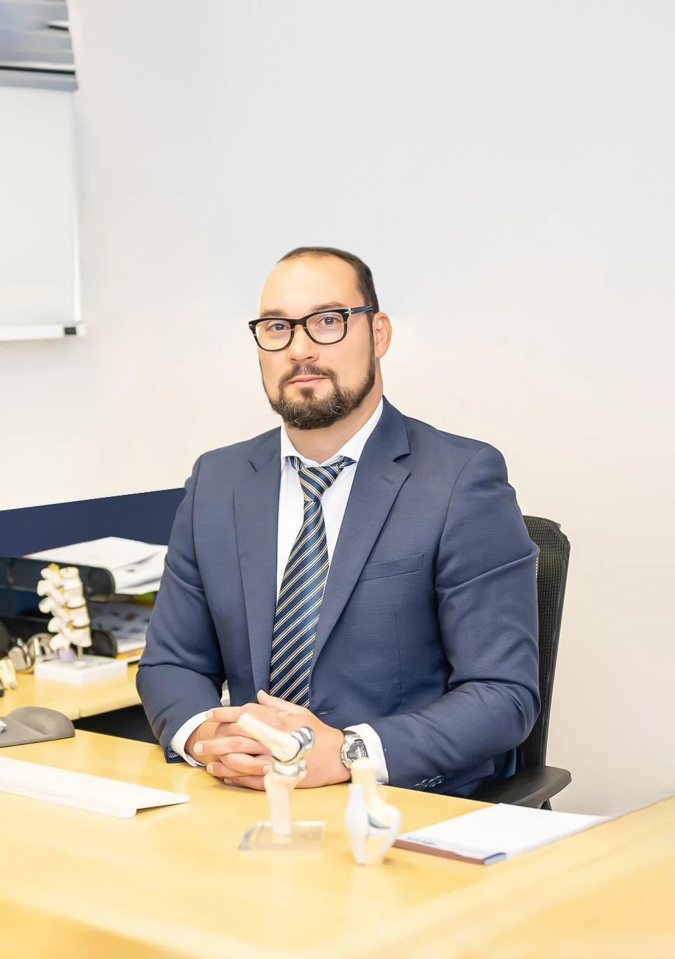 Hombre de negocios con gafas y barba, vestido con saco y corbata, sentado en una oficina con escritorio, documentos y equipo de oficina.
