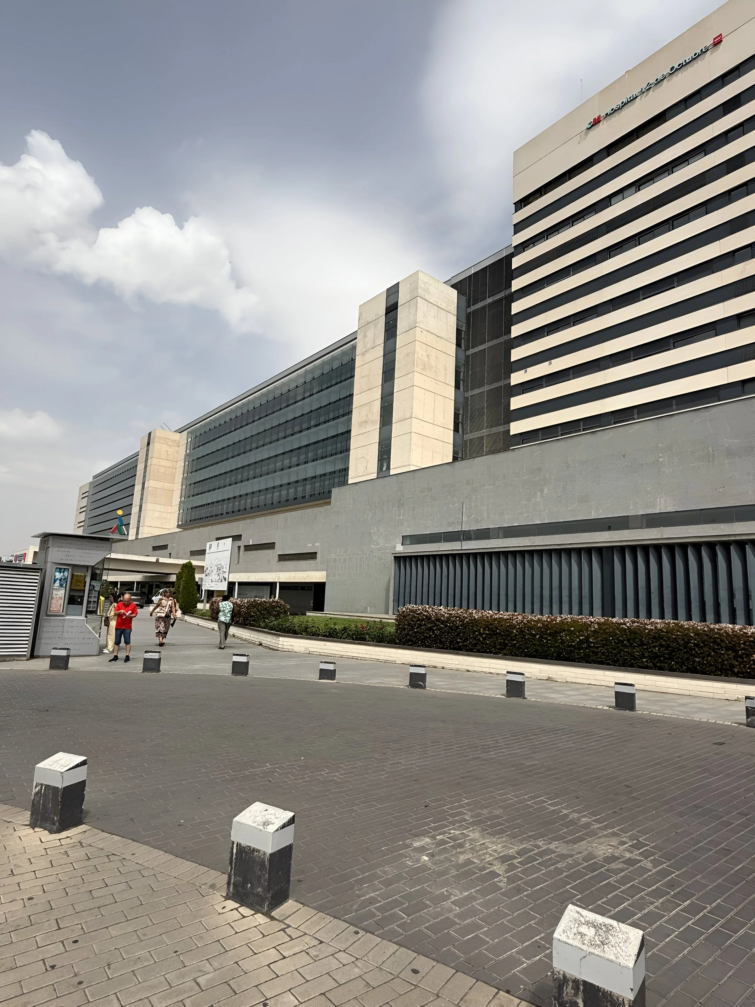 Edificio del Hospital Universitario 12 de Octubre en España, en un día soleado con algunas nubes y personas caminando afuera.