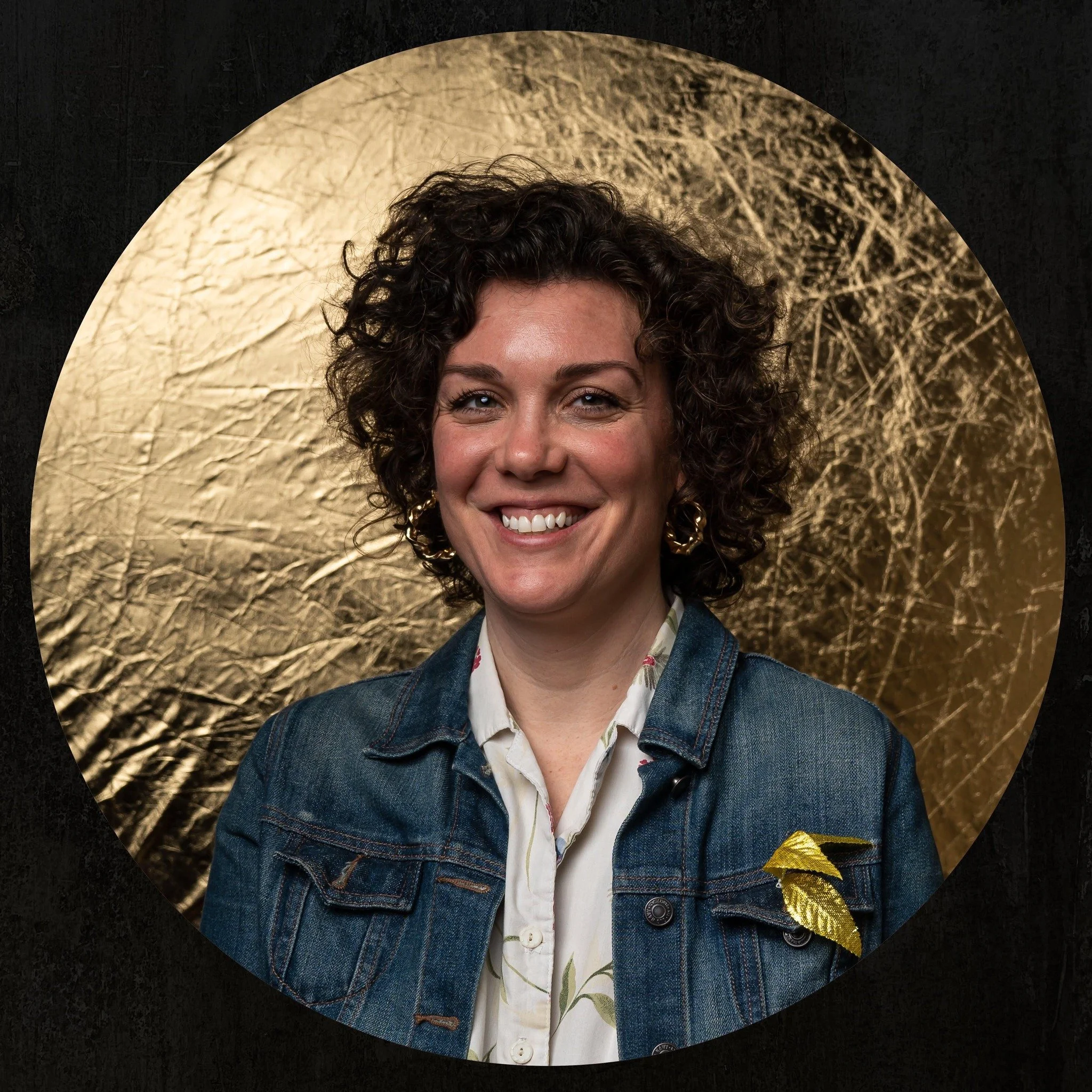 Portrait of a woman smiling, with short curly hair, wearing a denim jacket, gold earrings, and a white blouse with a floral pattern, standing in front of a textured gold background.