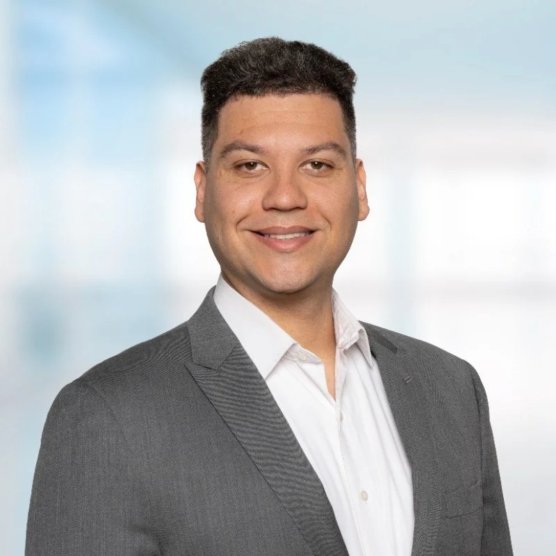 Professional headshot of a man wearing a gray suit and white shirt, smiling with a blurred office background.