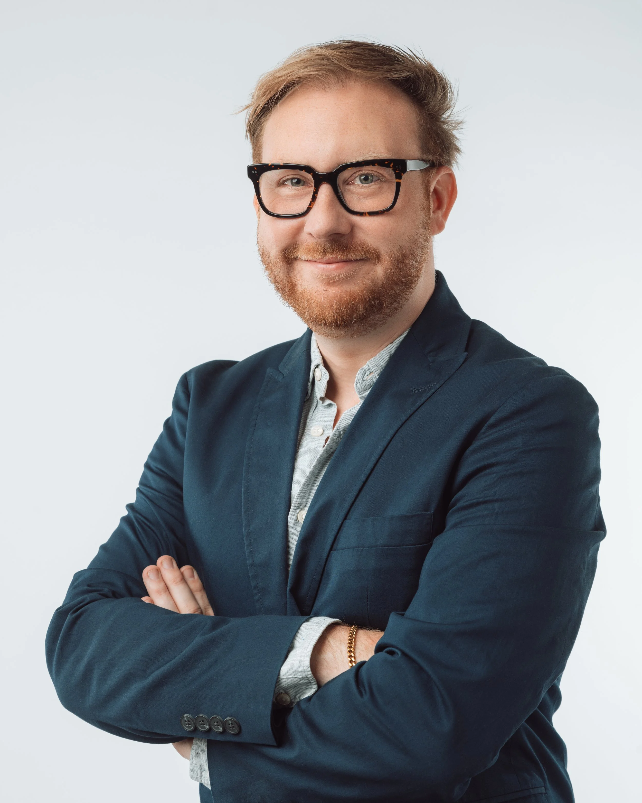 A man in glasses, a dark blazer, and a light shirt smiling with arms crossed against a plain background.