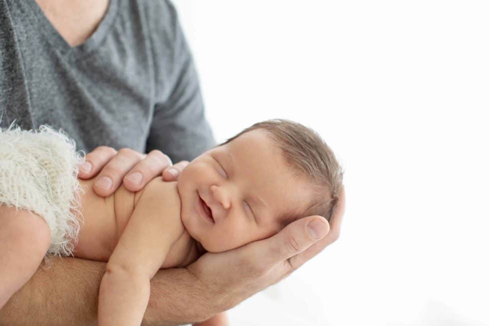 A person holds a sleeping newborn baby with a smile on their face, in a bright setting.