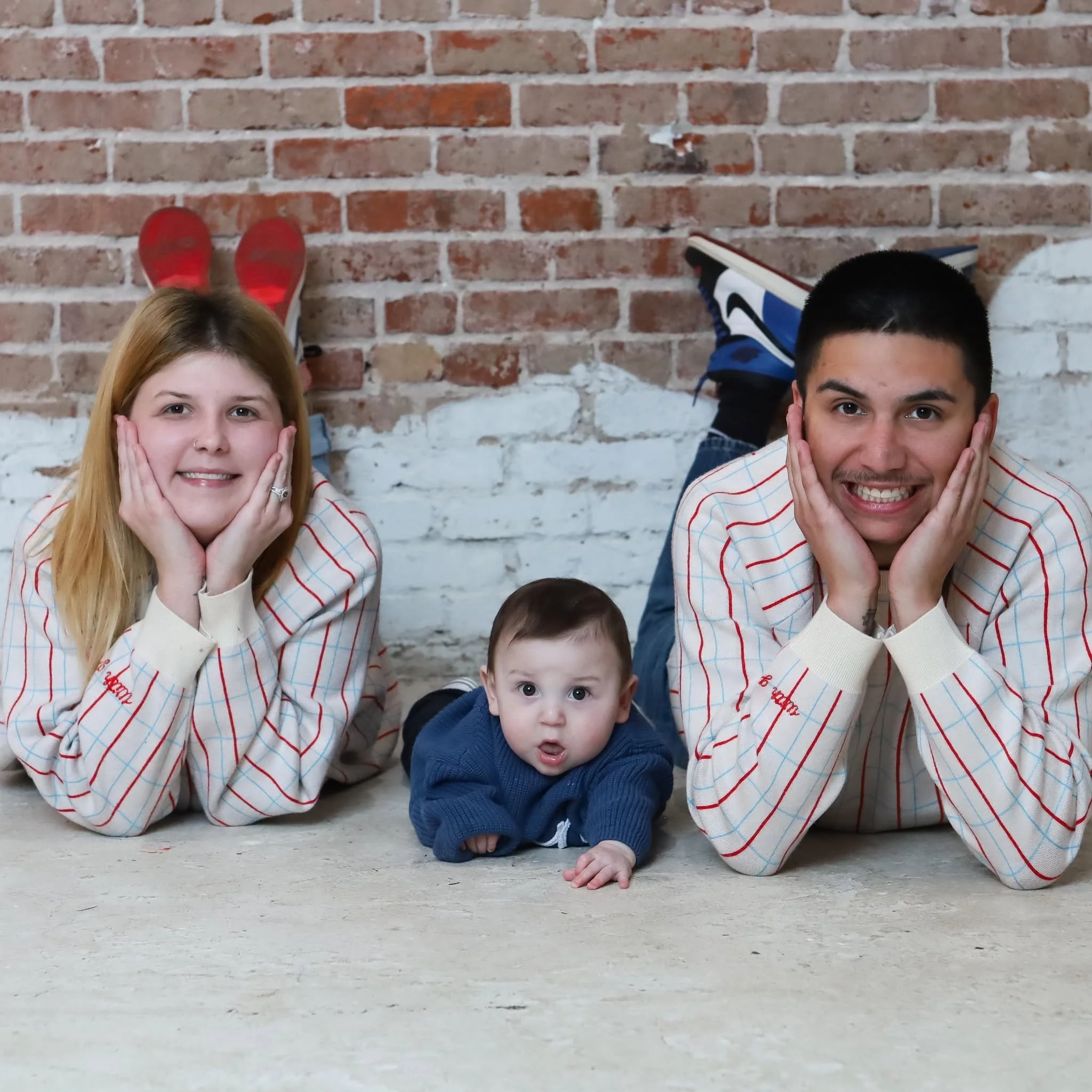Two children and a baby lying on the floor against a brick wall. The children are smiling and have their hands on their cheeks. One with red hair and a girl, the other with dark hair and a boy, all wearing light-colored shirts with red and blue stripes, and the baby has light brown hair and is dressed in blue.