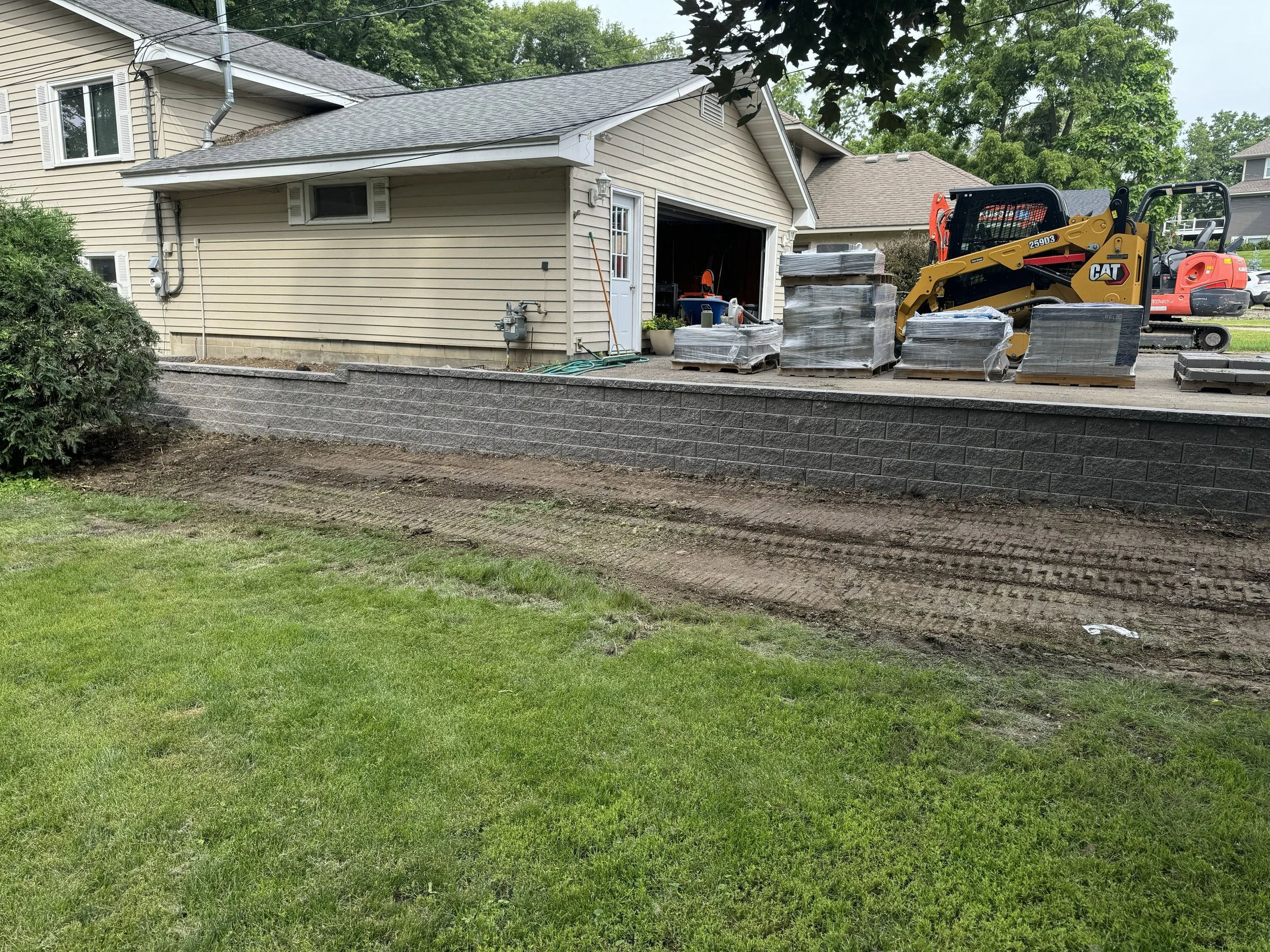 Backyard with a raised patio and stack of paving stones, construction equipment, and undisturbed grass in the foreground.