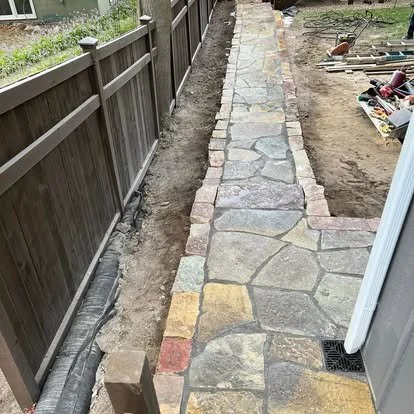 Stone walkway under construction next to a wooden fence and a house with tools and materials nearby.